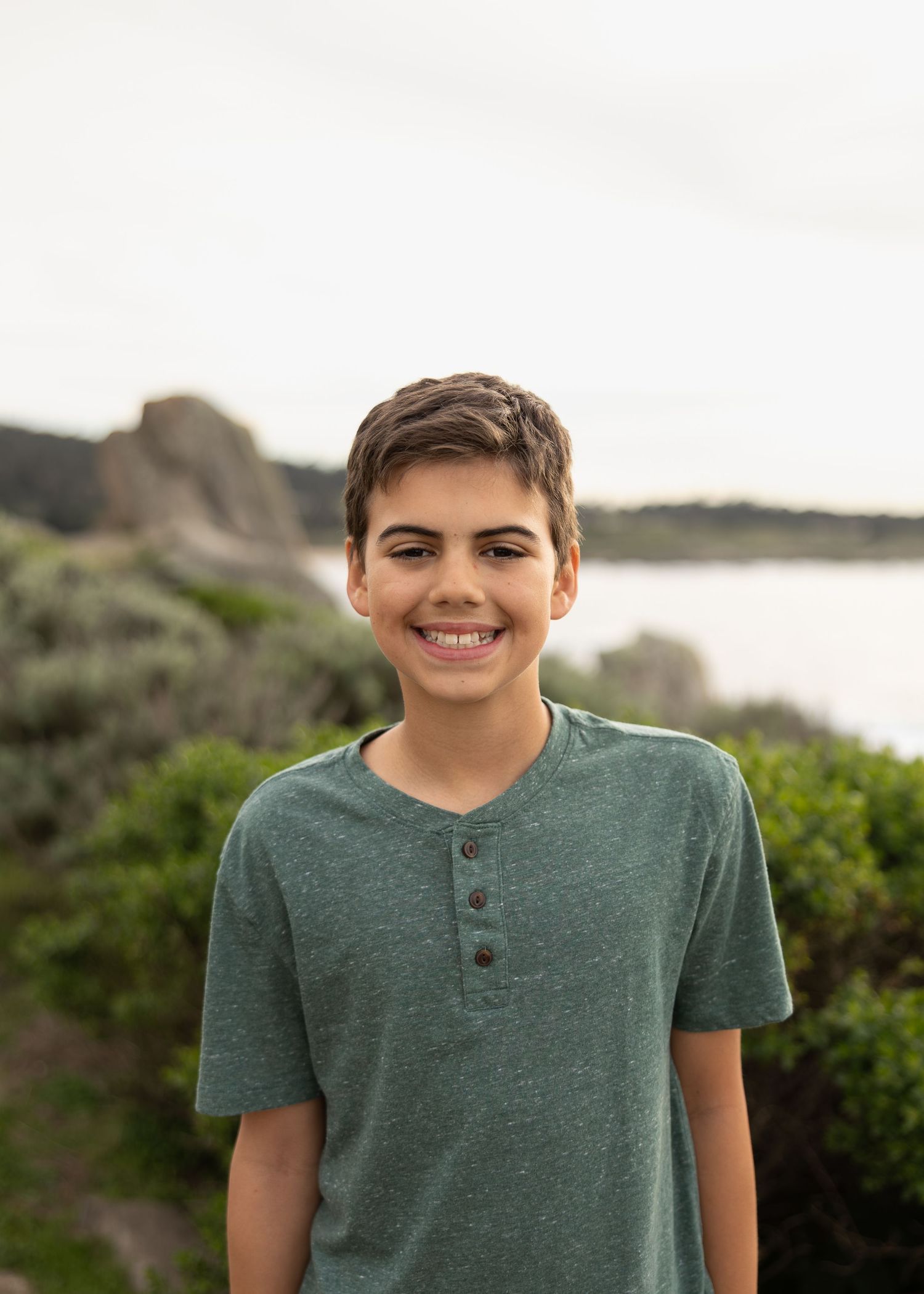 Happy young boy smiling outdoors in a green henley shirt with coastal rocks and shrubs in background.