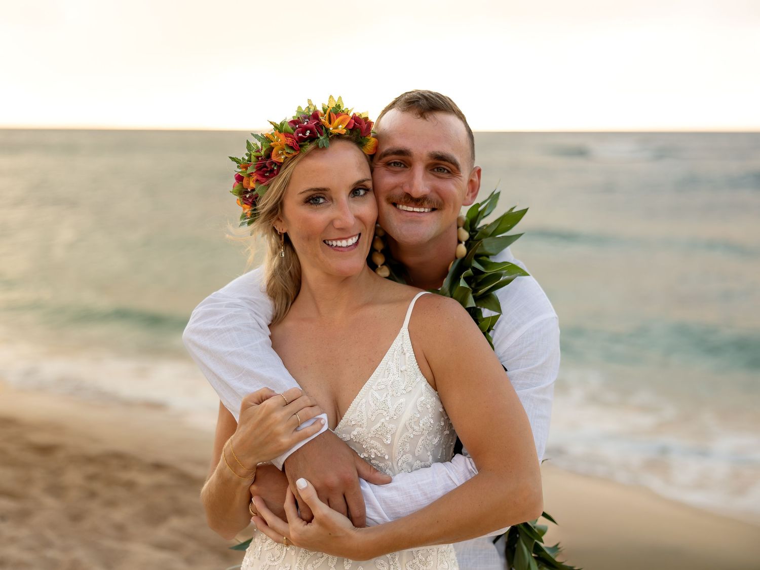 Couple embracing on a Hawaiian beach at sunset, bride in lace gown with floral crown, groom in white shirt with lei.