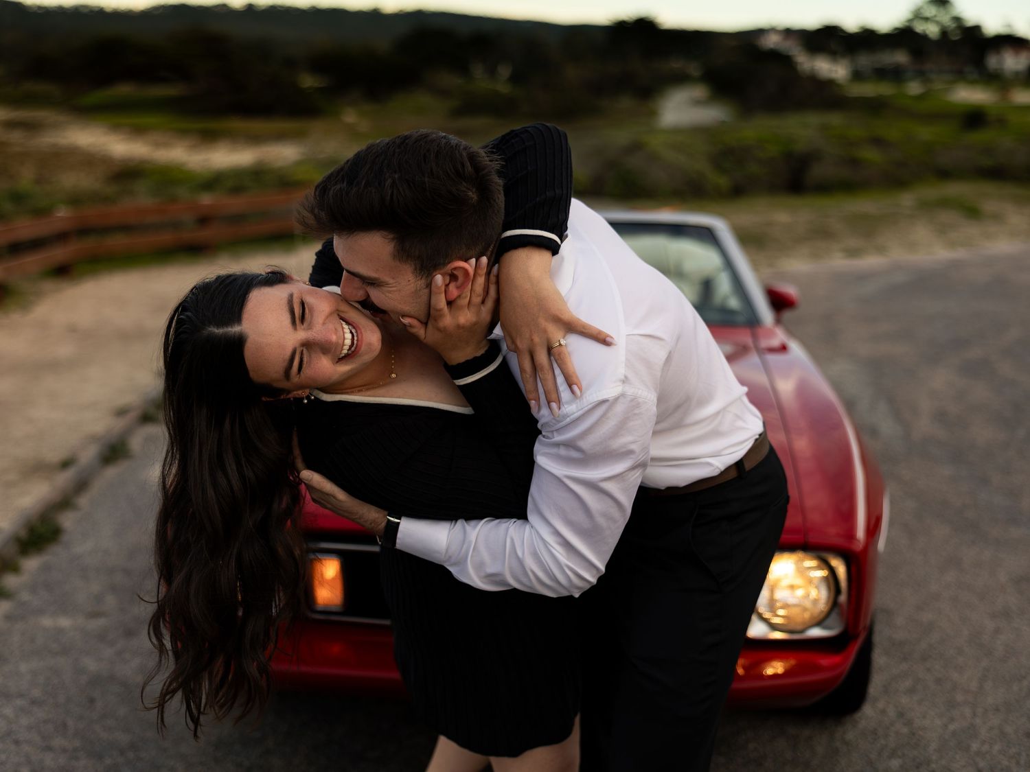 Couple embracing and laughing in front of a red convertible on a scenic road at dusk.