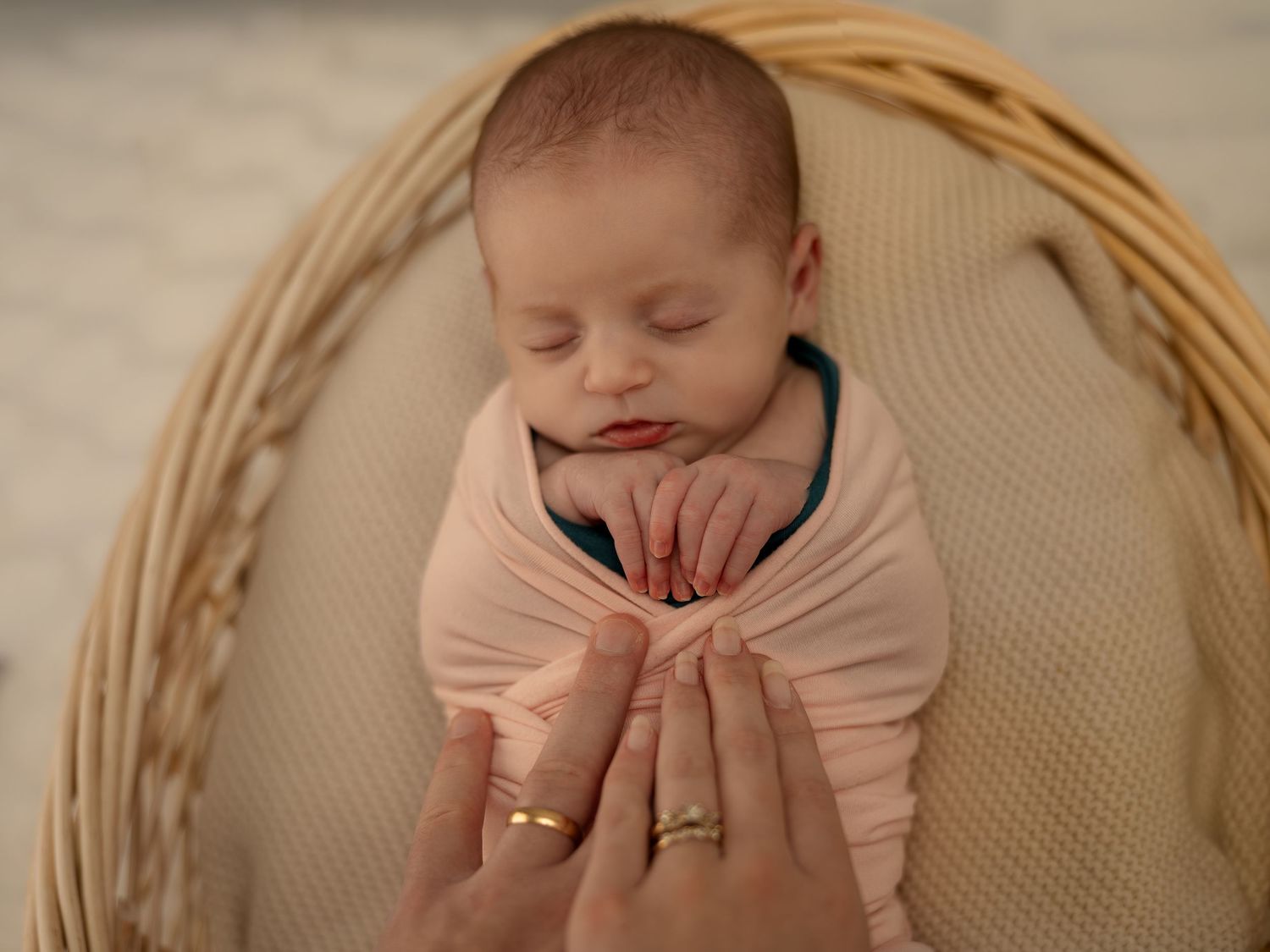 Newborn baby swaddled in pink wrap sleeping peacefully in wicker basket while parent gently holds tiny hands.