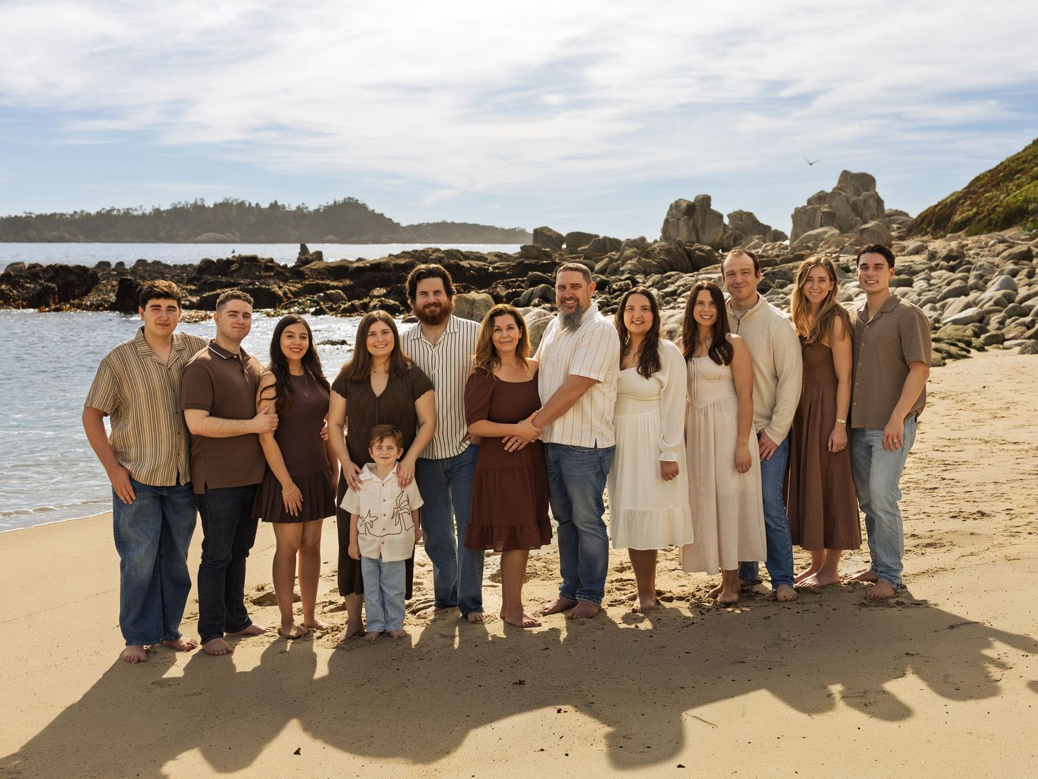 Large family group portrait on a sandy beach with rocky coastline backdrop on a cloudy day.