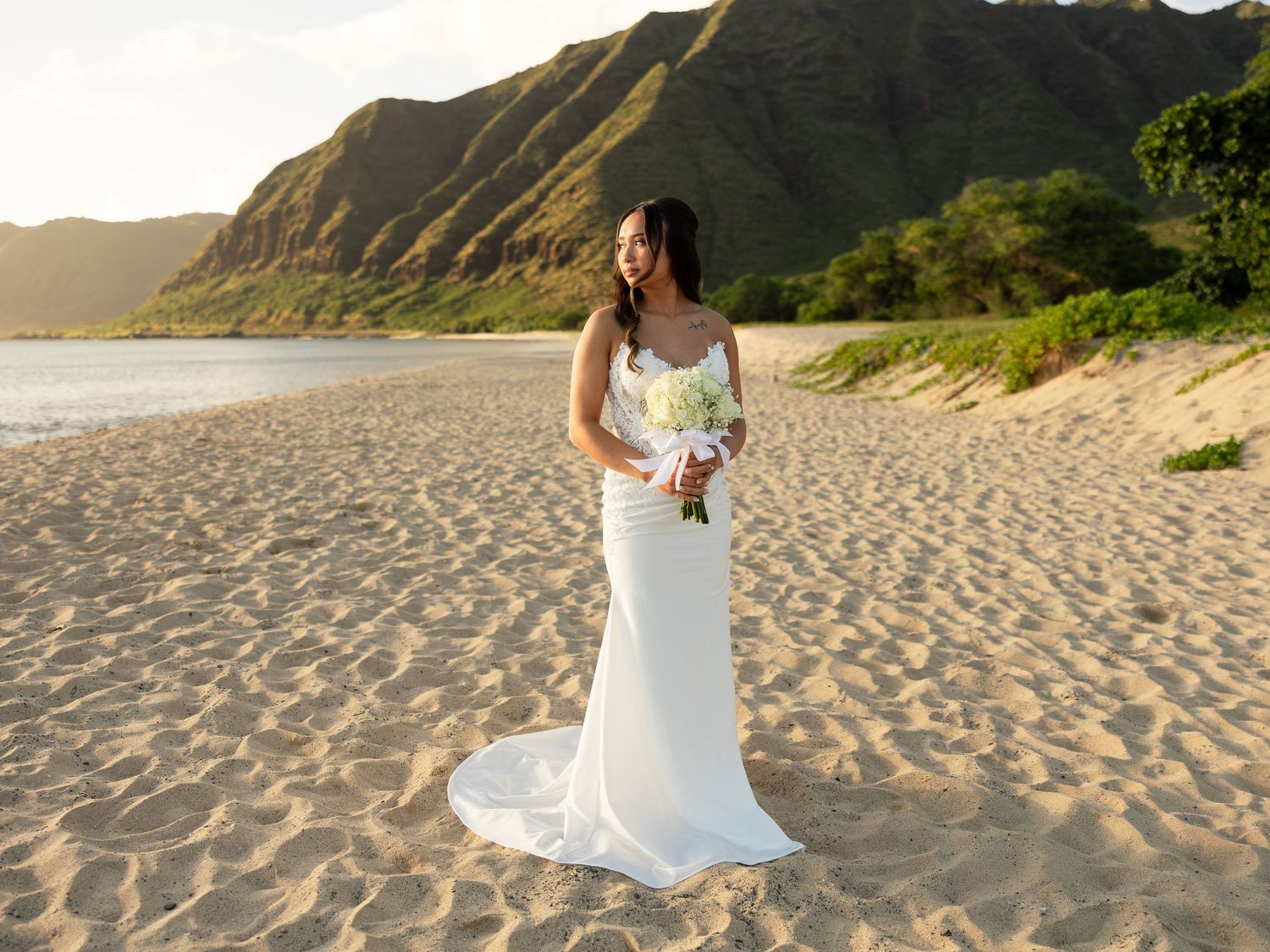 Bride in white gown holding bouquet on sandy beach with lush green mountains at sunset in Hawaii.