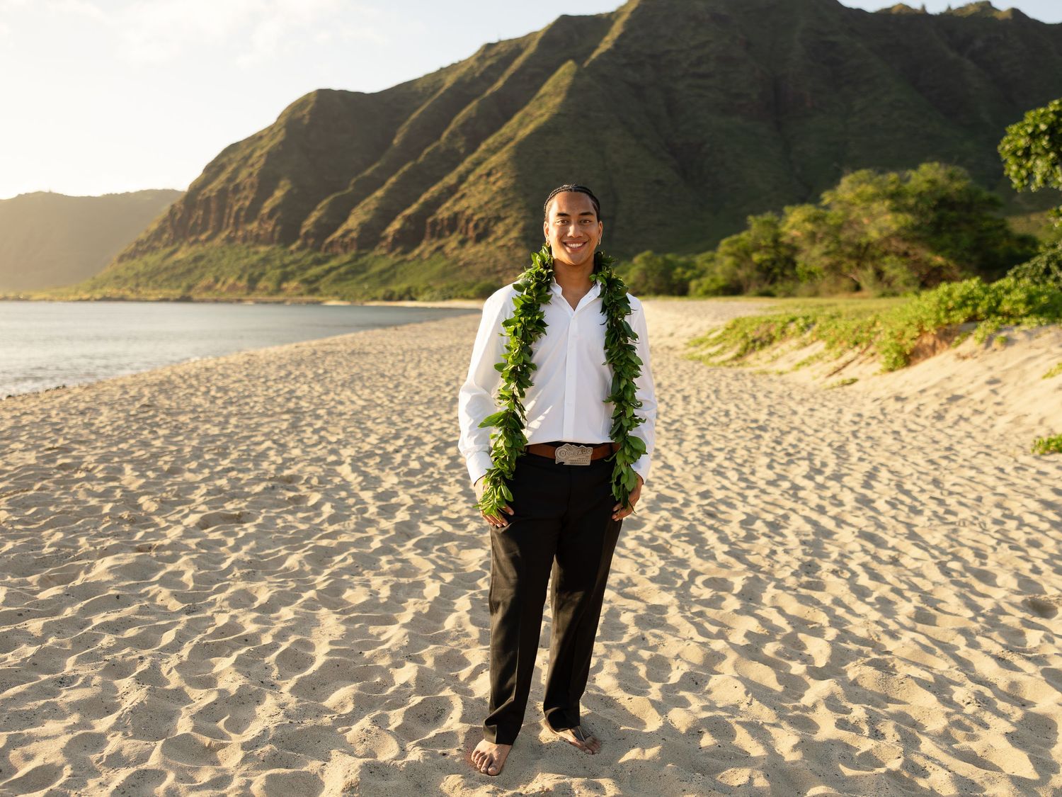 Person wearing lei standing barefoot on sandy Hawaiian beach with lush green mountains in golden sunset light.