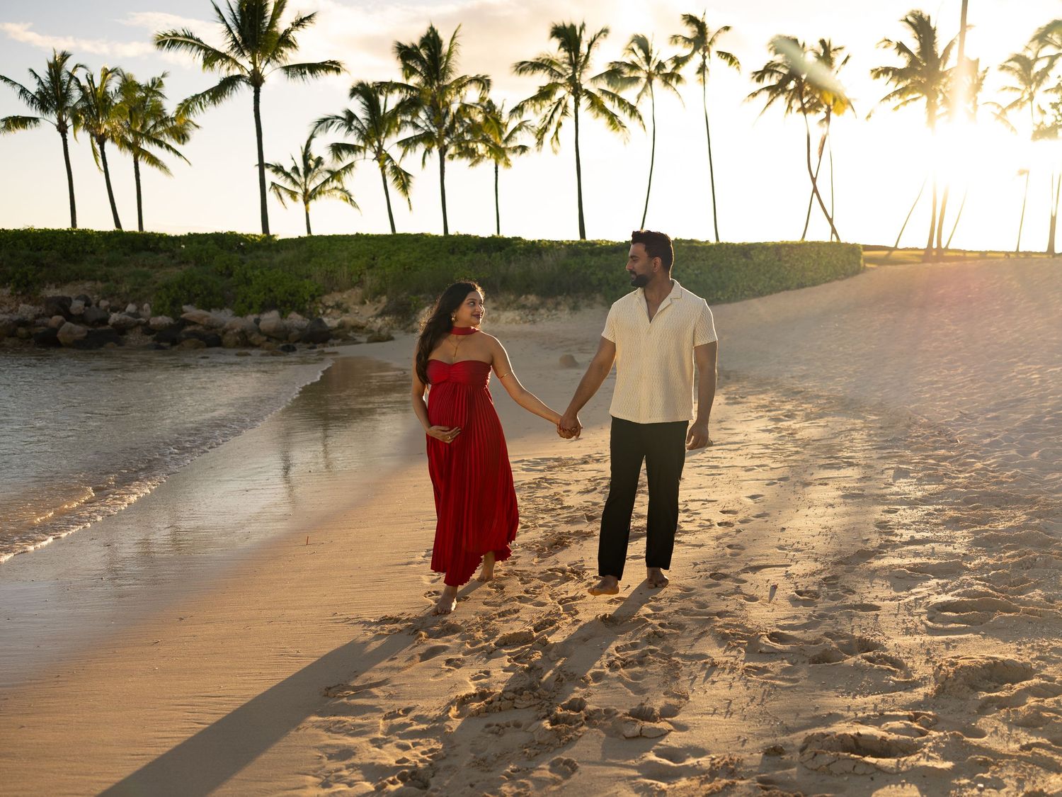 Pregnant couple holding hands on tropical beach at sunset, woman in red dress, palm trees in background.