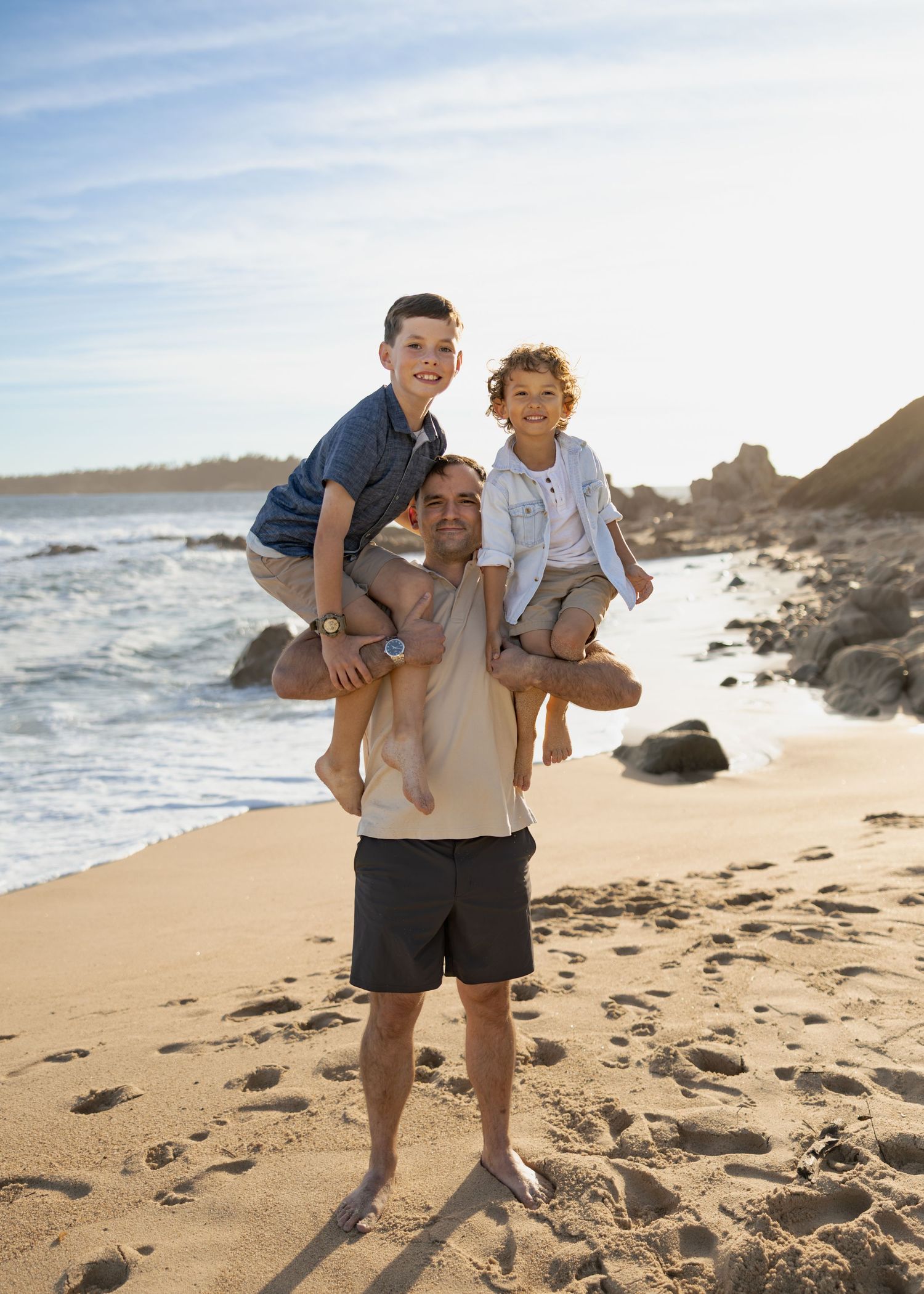 A father carries two young boys on his shoulders while smiling on a sunny sandy beach with ocean waves behind them.