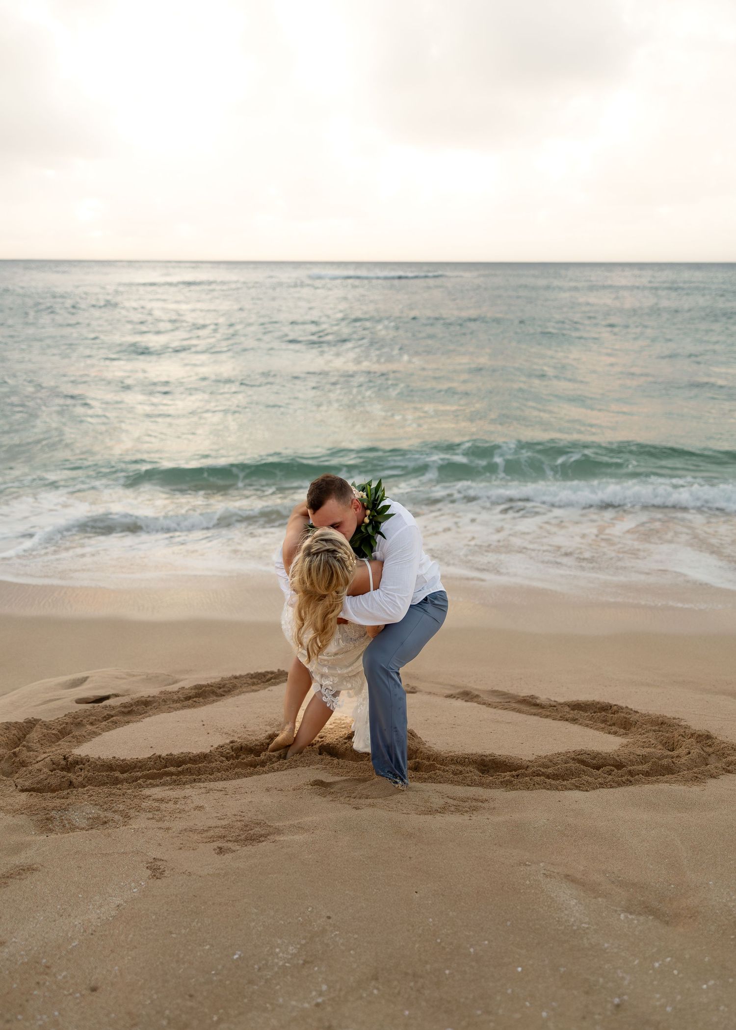 Bride and groom sharing a romantic kiss on a sandy beach with a heart drawn in the sand and ocean waves behind them.