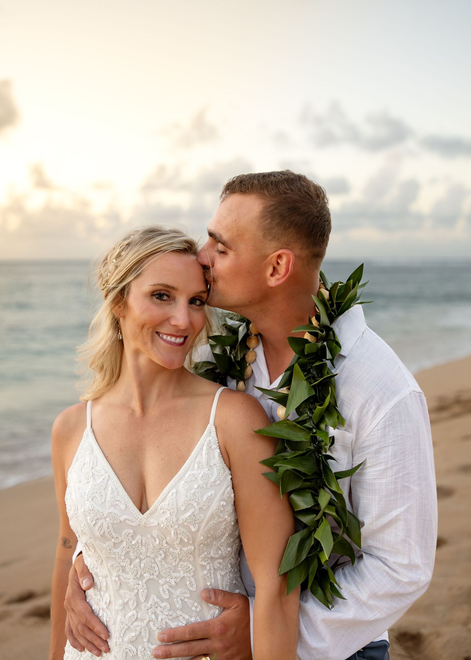 Couple sharing a romantic beach sunset kiss, bride in white lace gown, groom wearing a green leaf lei in Hawaii.