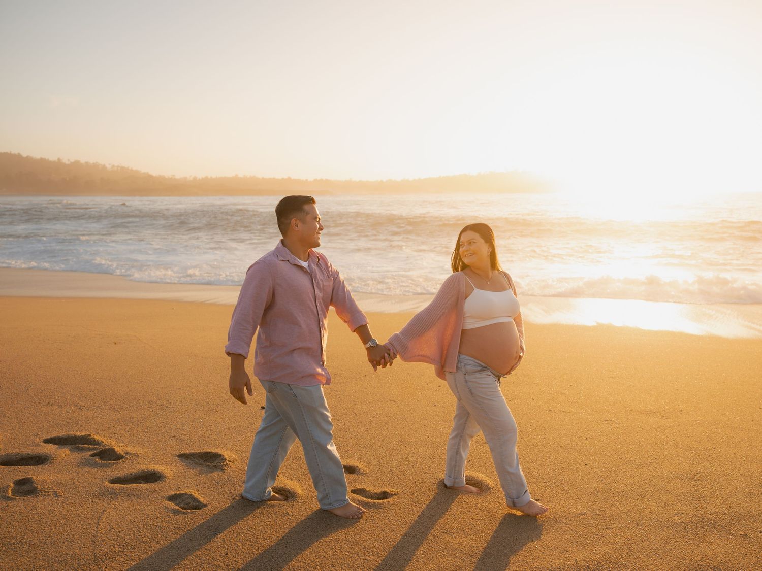 Pregnant couple walks hand-in-hand on a sunlit beach at golden hour with ocean waves behind them.