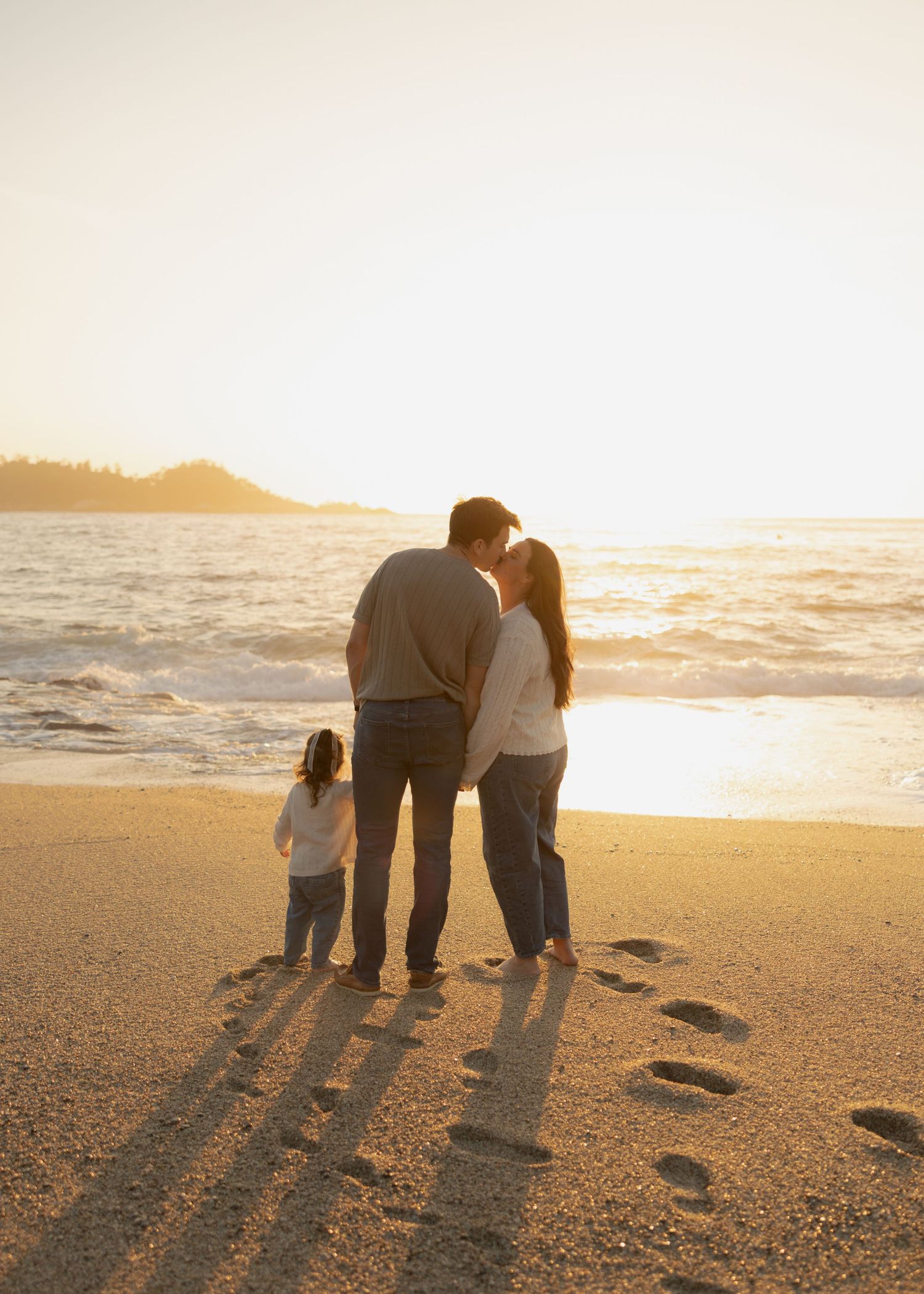 Family of three standing on a sandy beach at sunset, with footprints in the sand and ocean waves in the background.