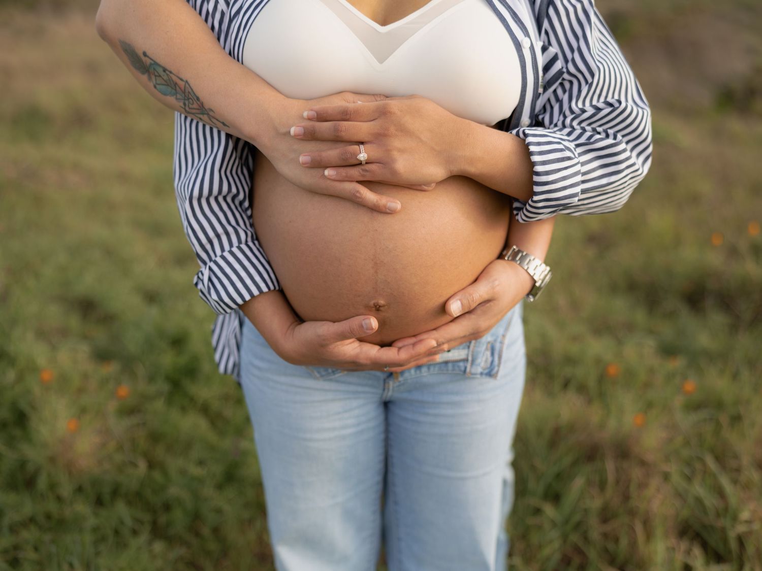 Close-up of pregnant woman cradling her bare baby bump outdoors in a field wearing jeans and striped shirt.