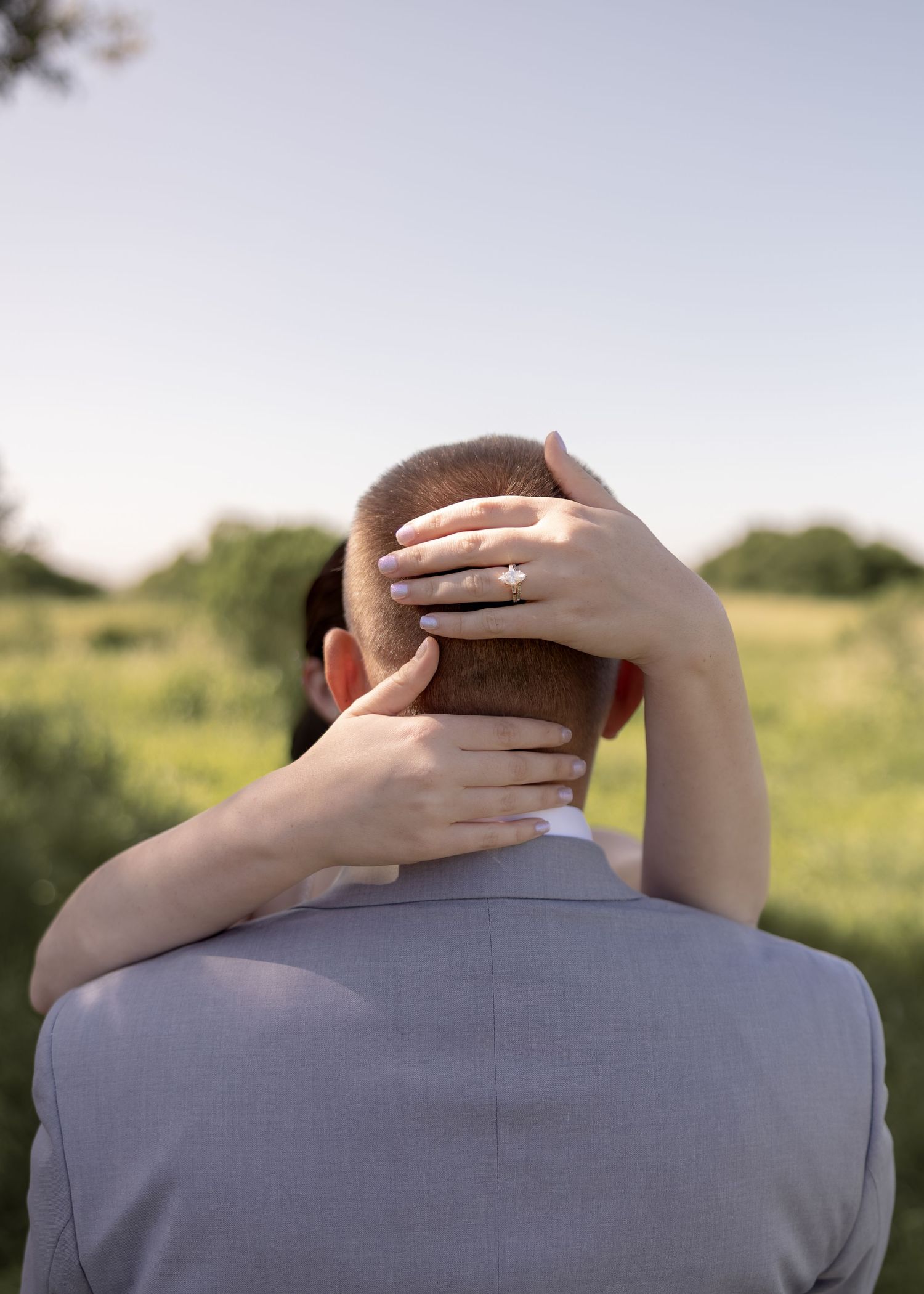A woman with an engagement ring rests her hand on a man's head outdoors in a green field.