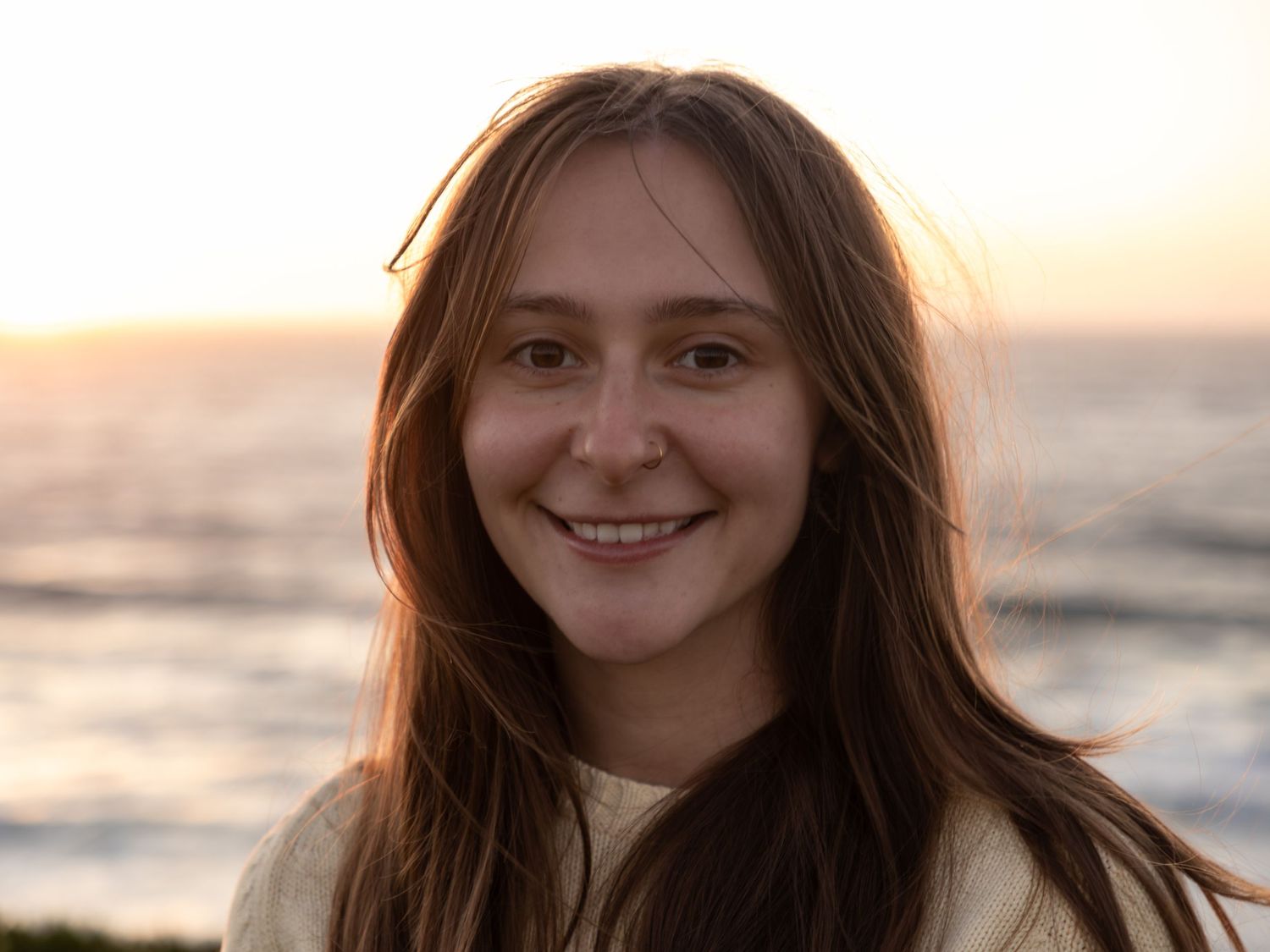 Smiling young woman with long brown hair at sunset beach, golden hour glow behind her.