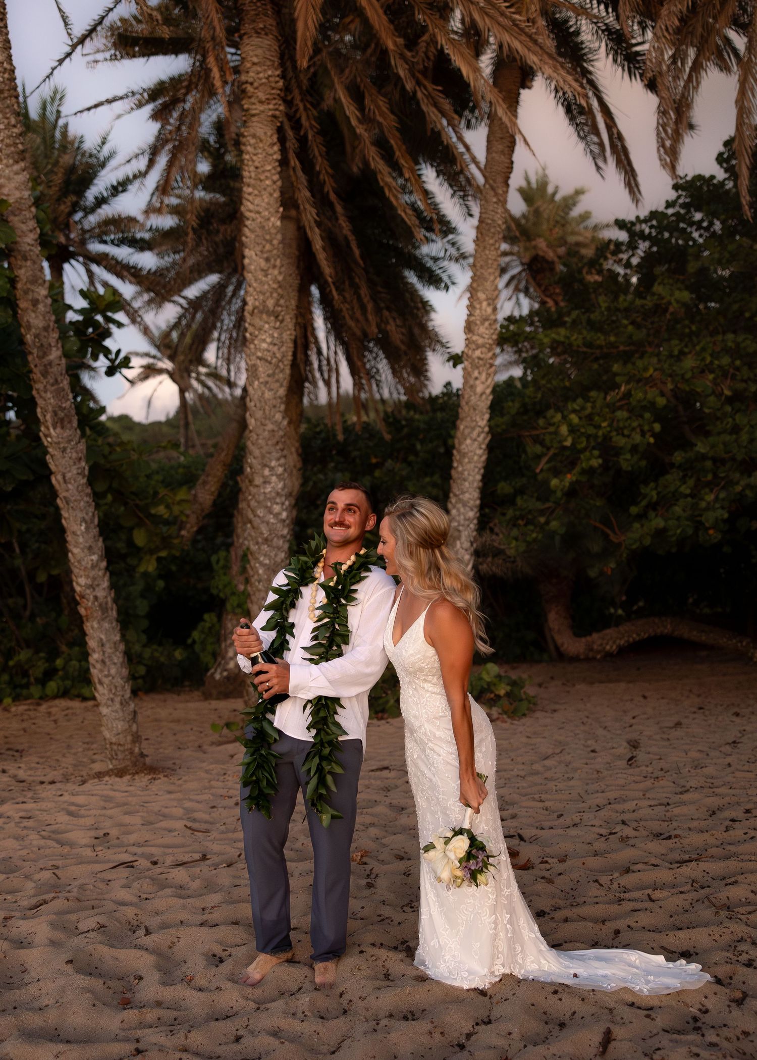 Couple in wedding attire standing on sandy beach with palm trees, groom in gray pants with lei, bride in white gown holding bouquet.