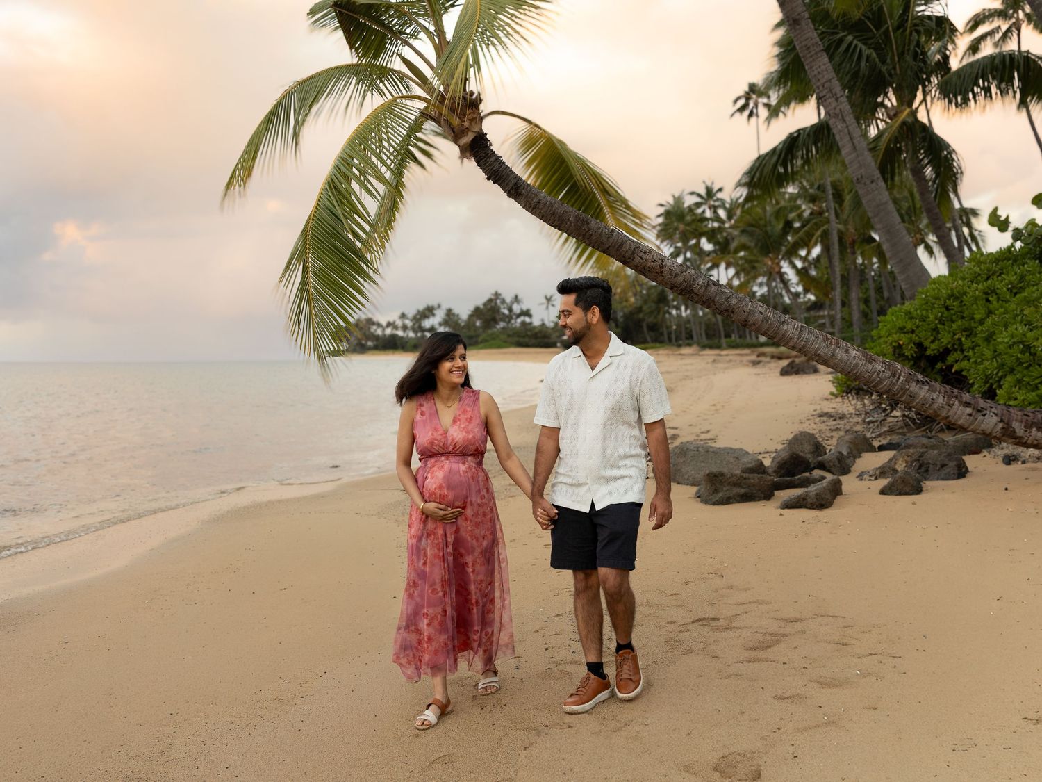 Pregnant couple walking hand in hand on a tropical beach at sunset with palm trees and golden sand.