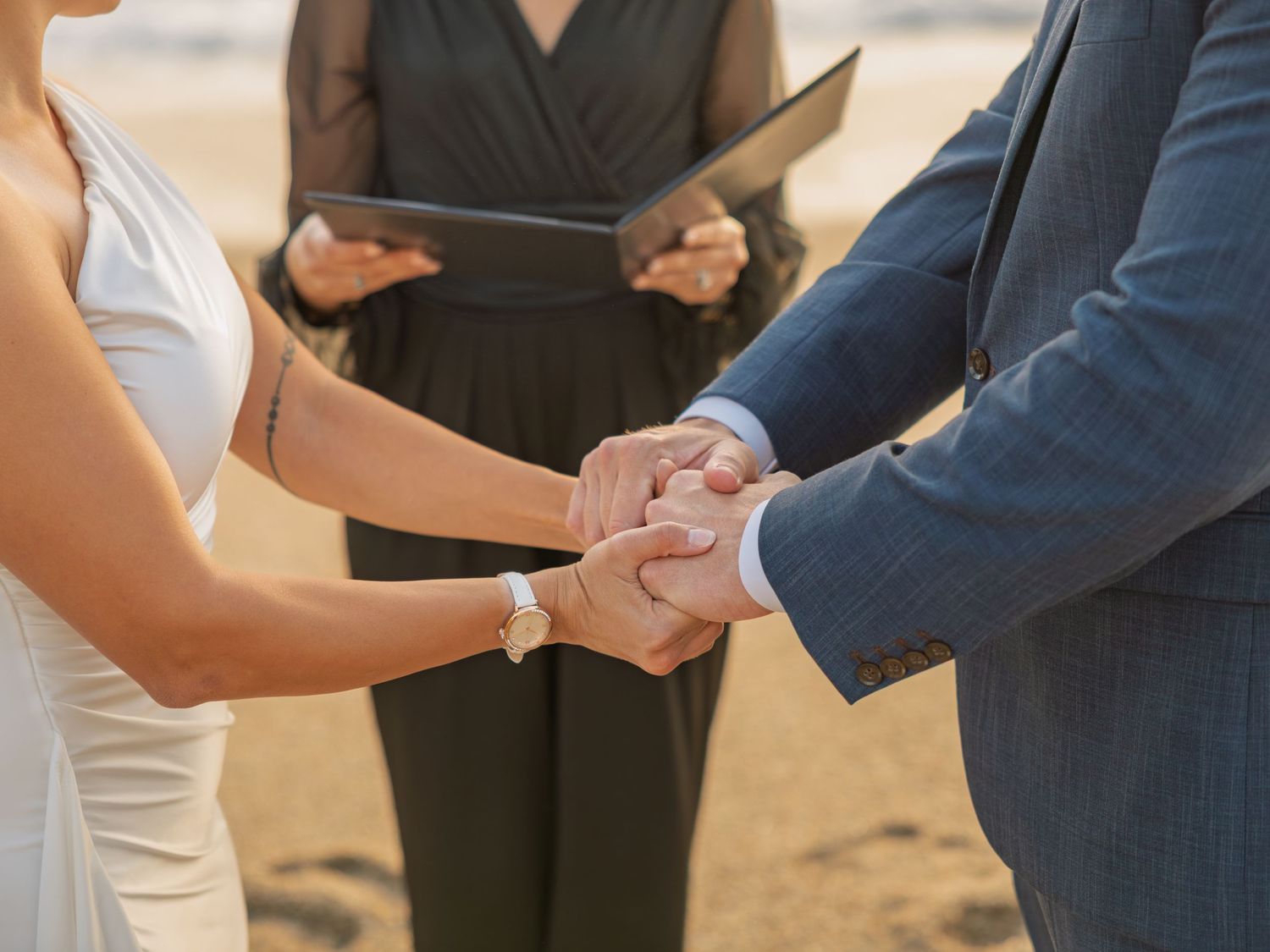 Close-up of bride and groom holding hands during outdoor beach wedding ceremony with officiant in background.