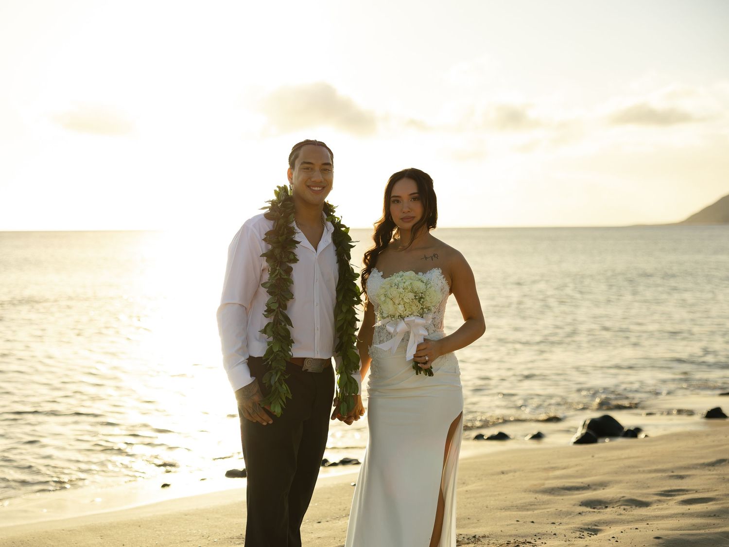 Couple in wedding attire posing on a beach at sunset, man wearing lei, woman holding white bouquet.