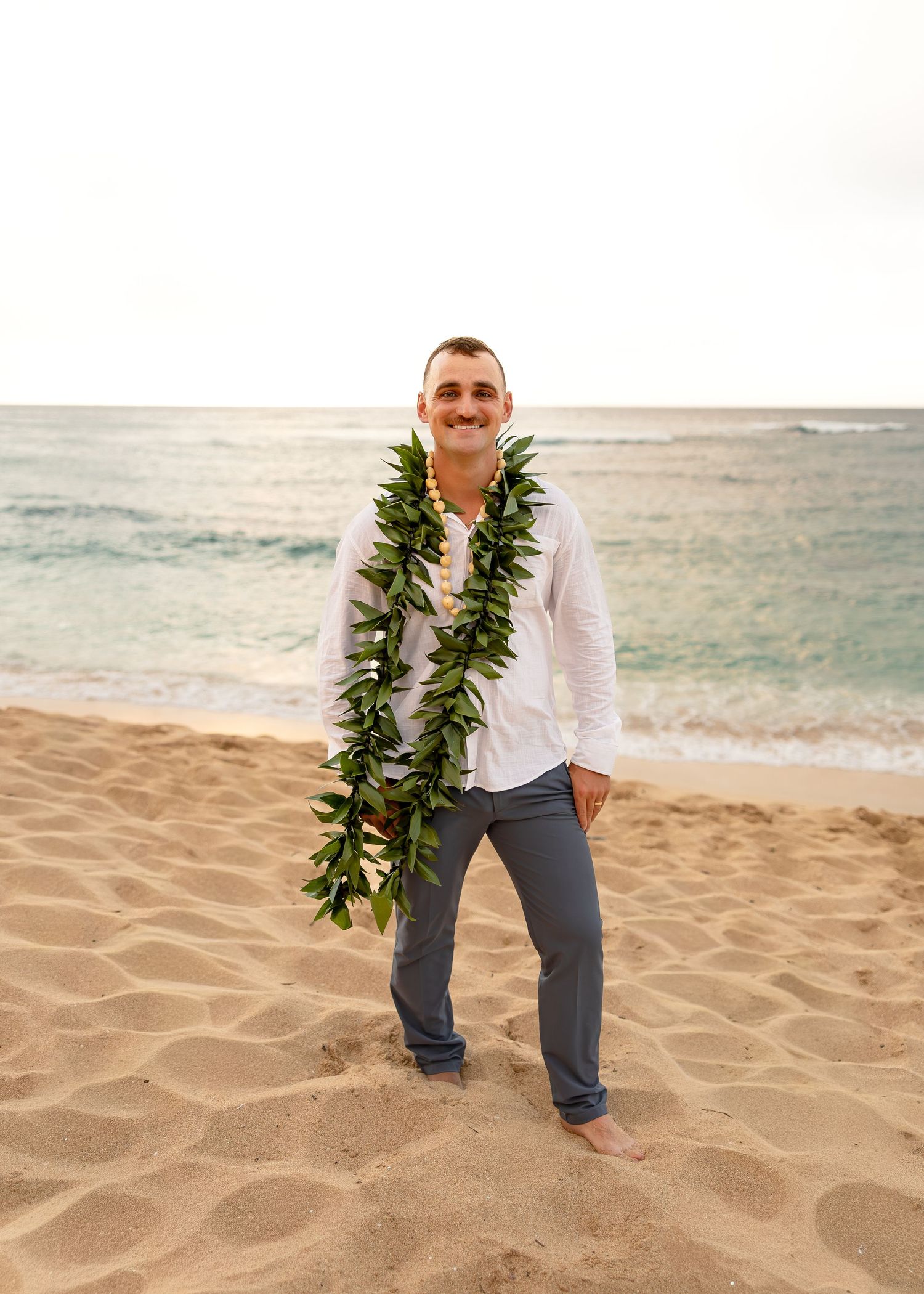 Smiling man wearing a green leaf lei and white shirt standing on a sandy Hawaiian beach with ocean waves behind him.