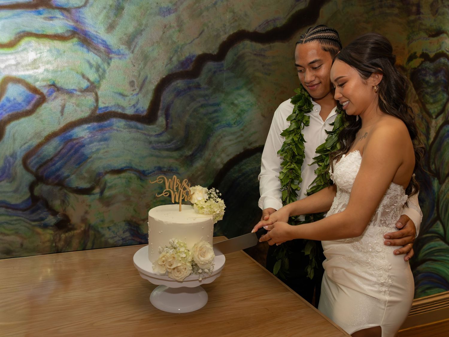 Couple in wedding attire cutting a white floral cake, wearing green lei garlands, with a colorful abstract background.