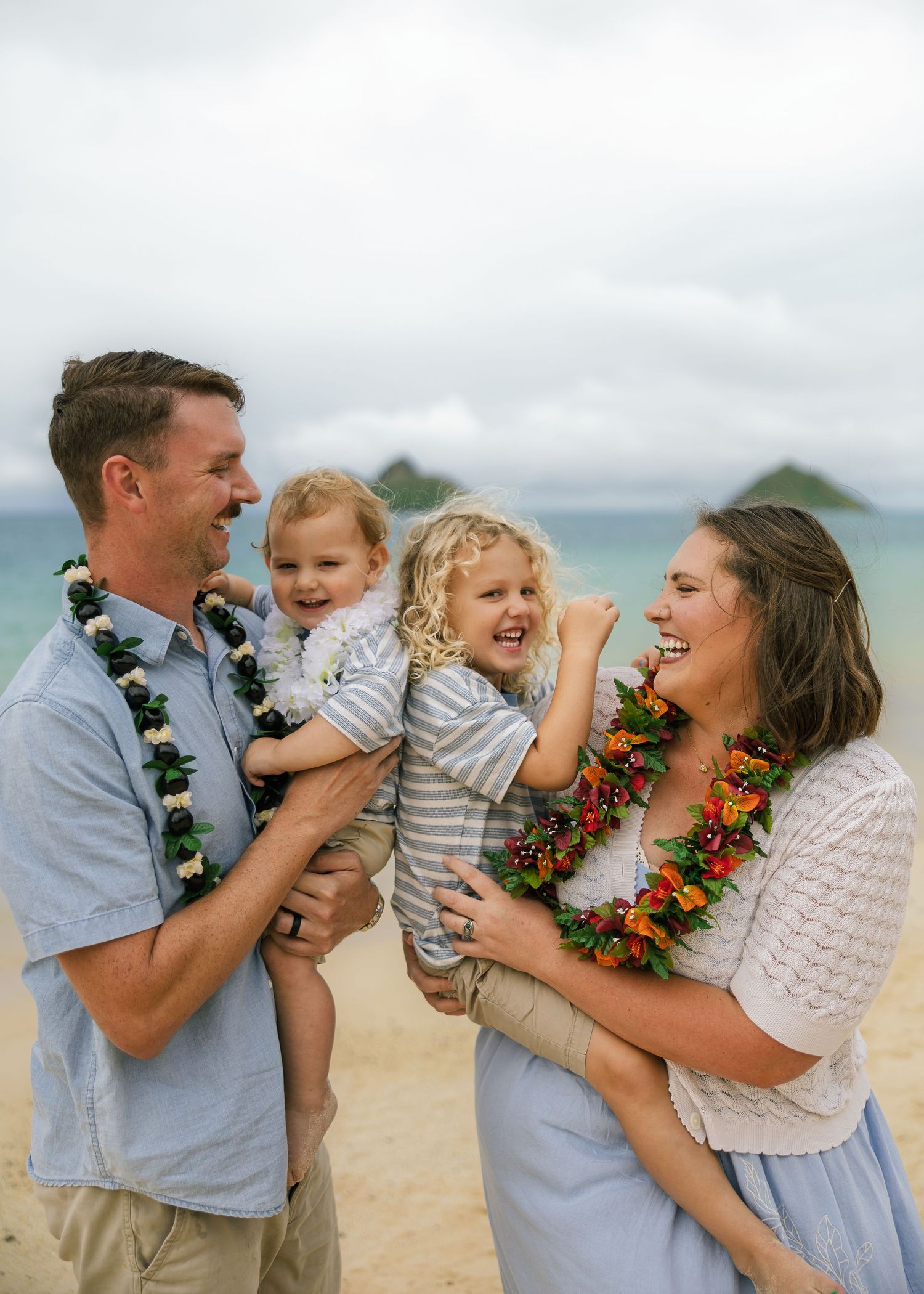 Family of four laughing together on a Hawaiian beach, wearing leis, with ocean and islands in the background.