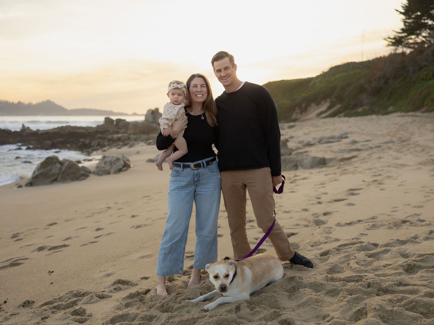 Family with baby and dog posing on sandy beach at sunset with ocean waves and green cliffs in background.