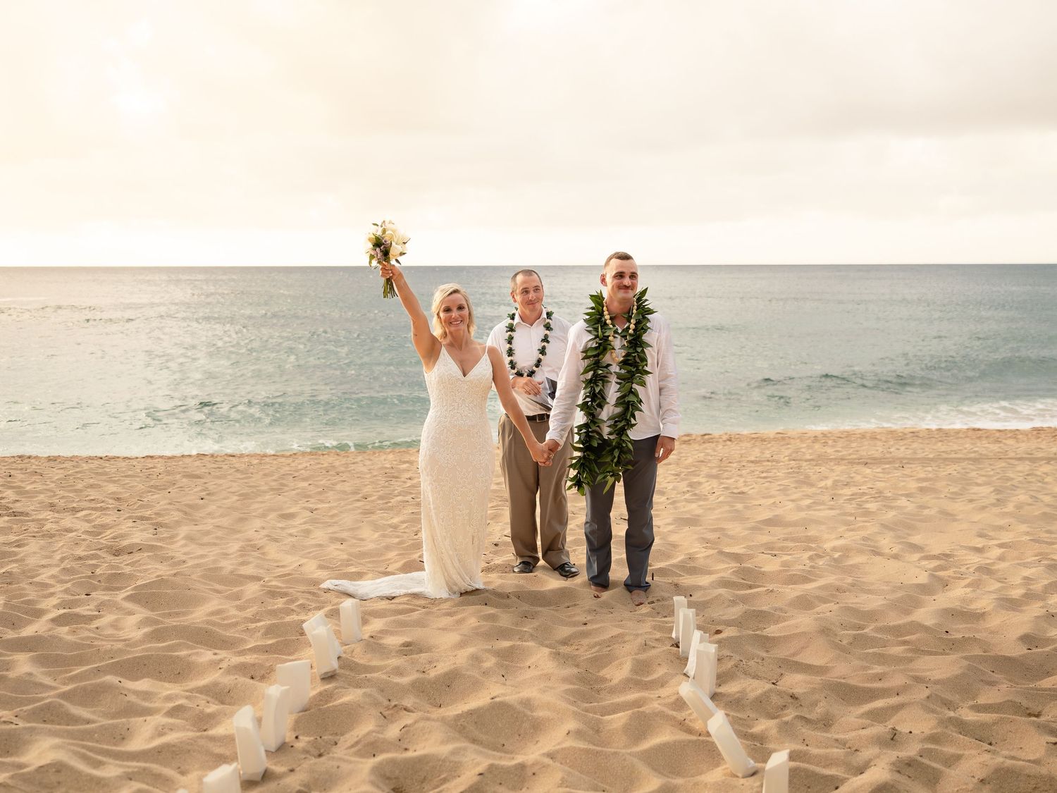 Couple celebrating beach wedding in Hawaii with officiant, bride in white dress raising bouquet, groom in lei, ocean backdrop.