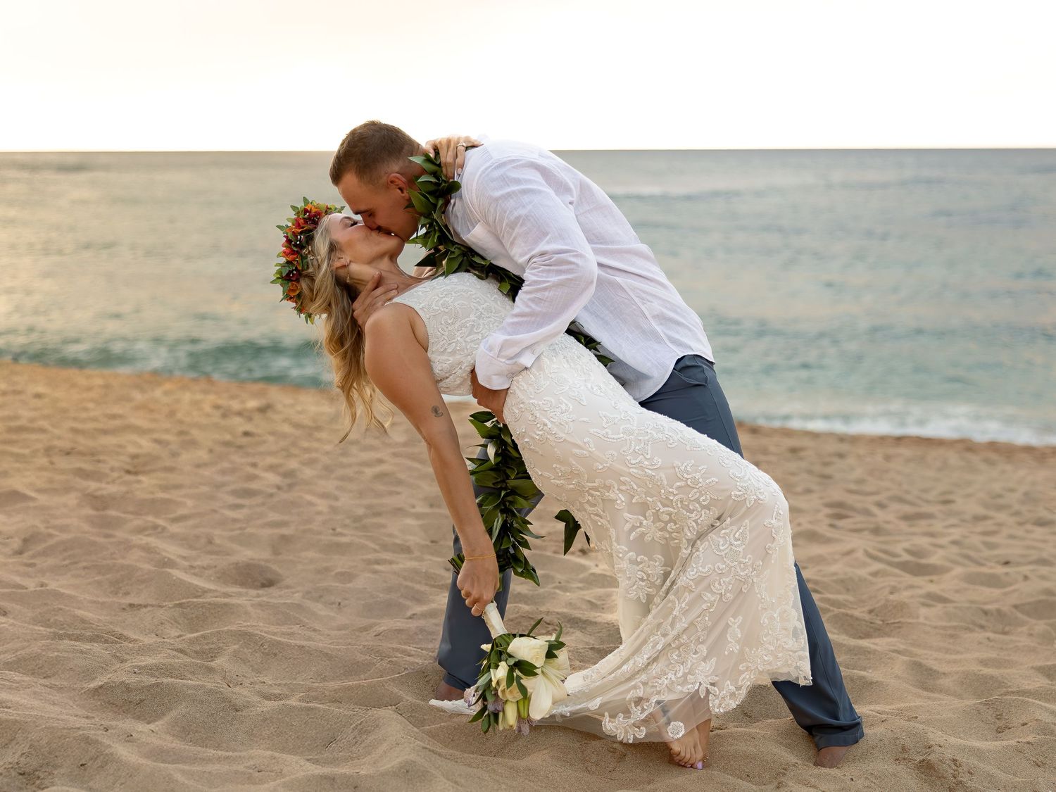 Couple sharing a romantic dip kiss on sandy beach, bride in lace gown with bouquet, groom in white shirt and jeans.