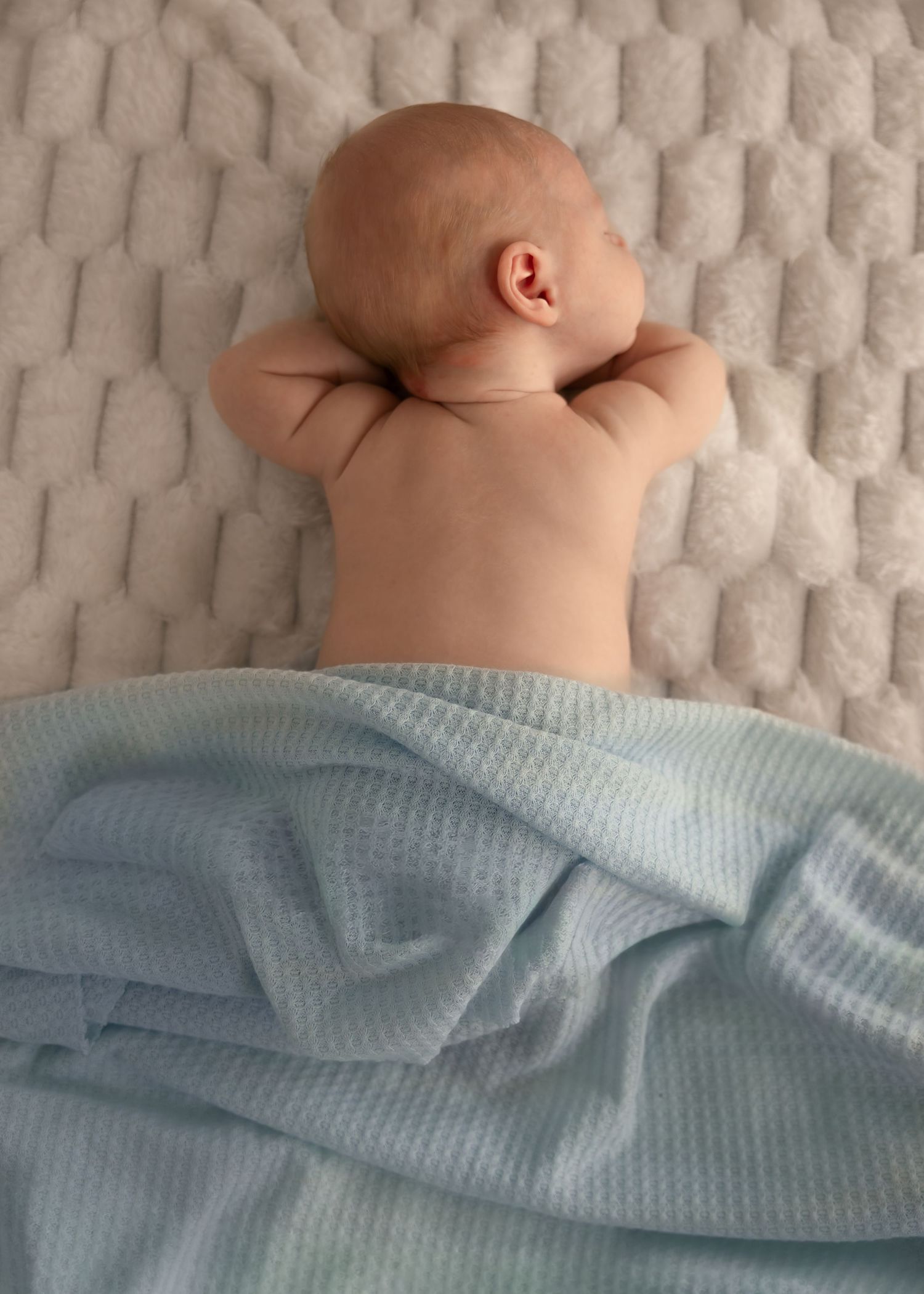 Newborn baby lying face down on a textured white mattress, covered with a soft light blue waffle blanket.