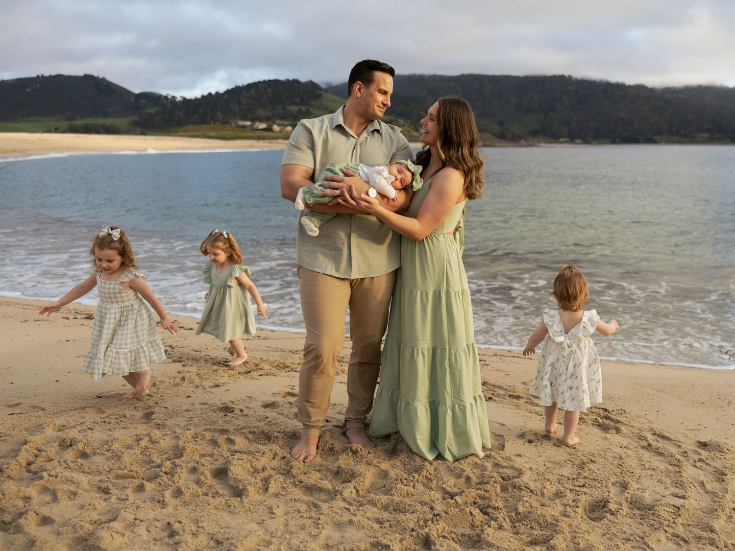 Family beach portrait with parents holding newborn and two young daughters in matching dresses on sandy shore.