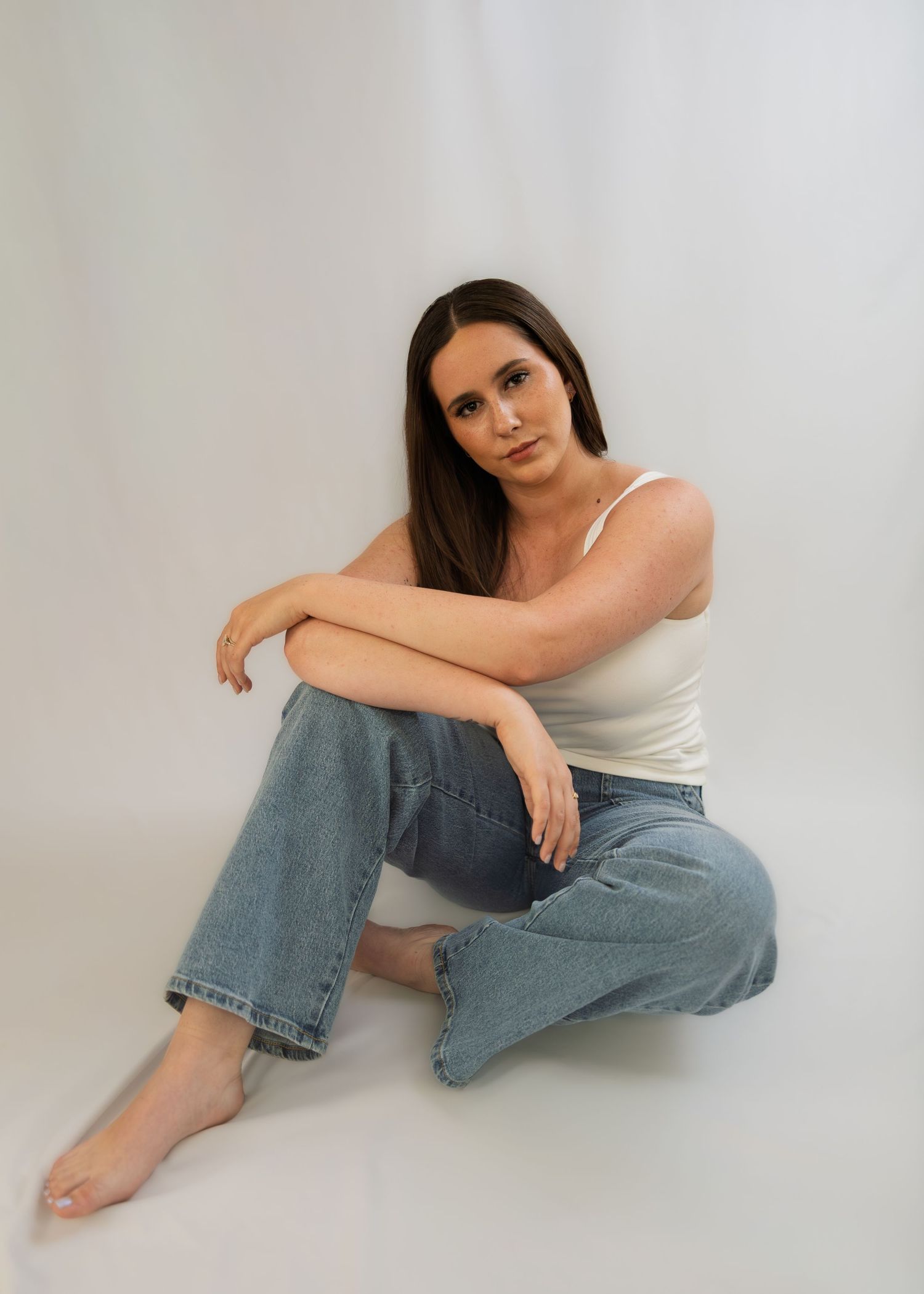 Young woman with long brown hair sitting on floor wearing white tank top and blue wide-leg jeans, barefoot.