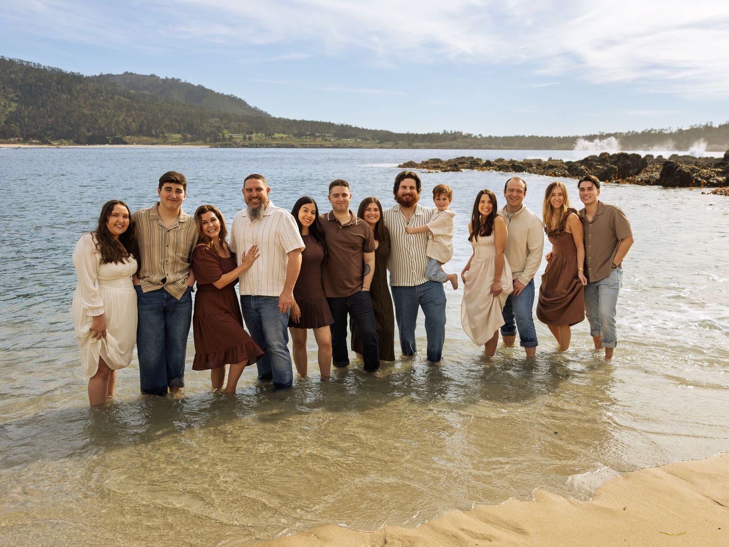 Large family group smiling together while standing in shallow ocean water on a sunny beach.