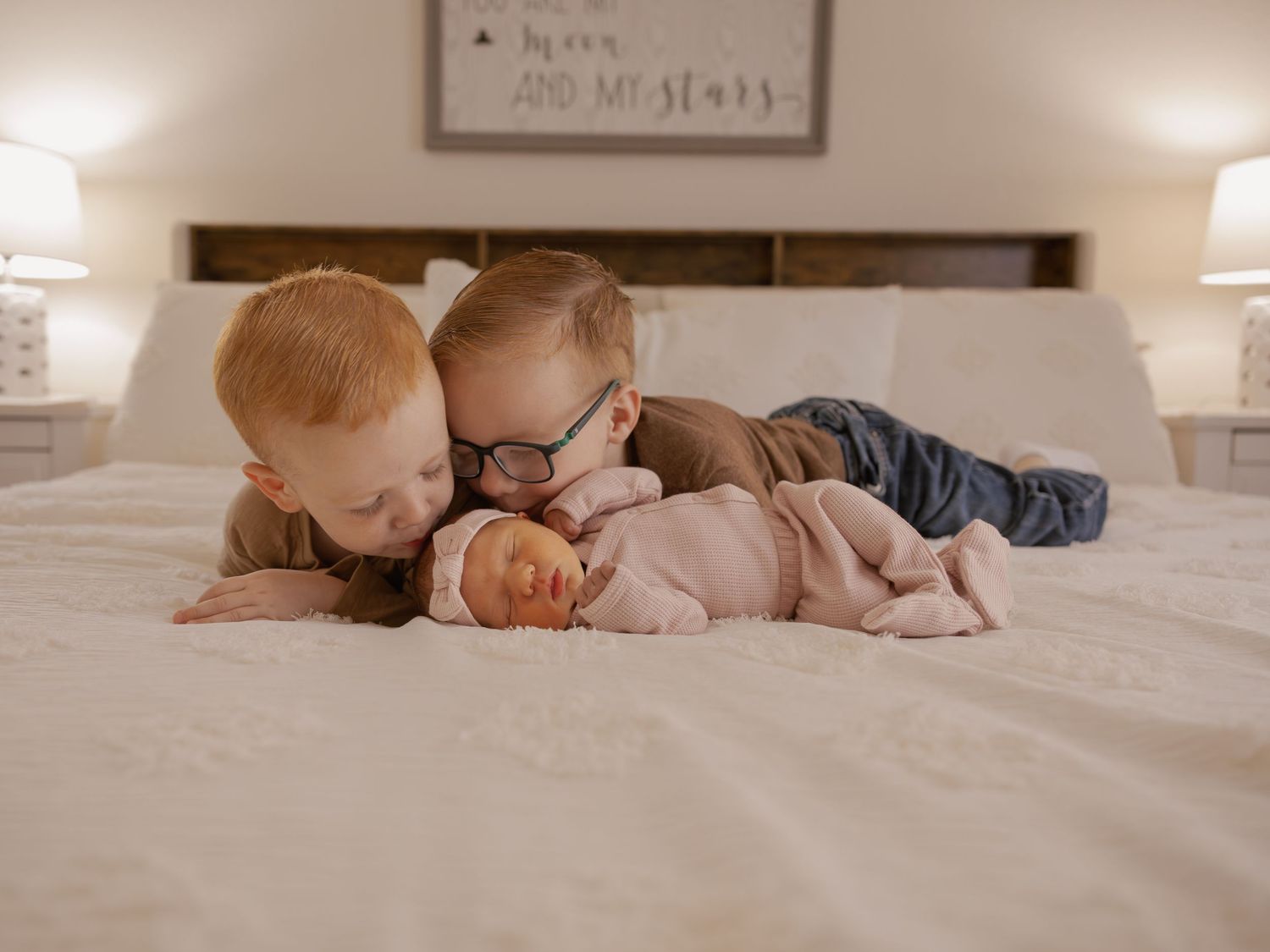 Two red-haired brothers lying on white bed gently kissing newborn baby sister between them.