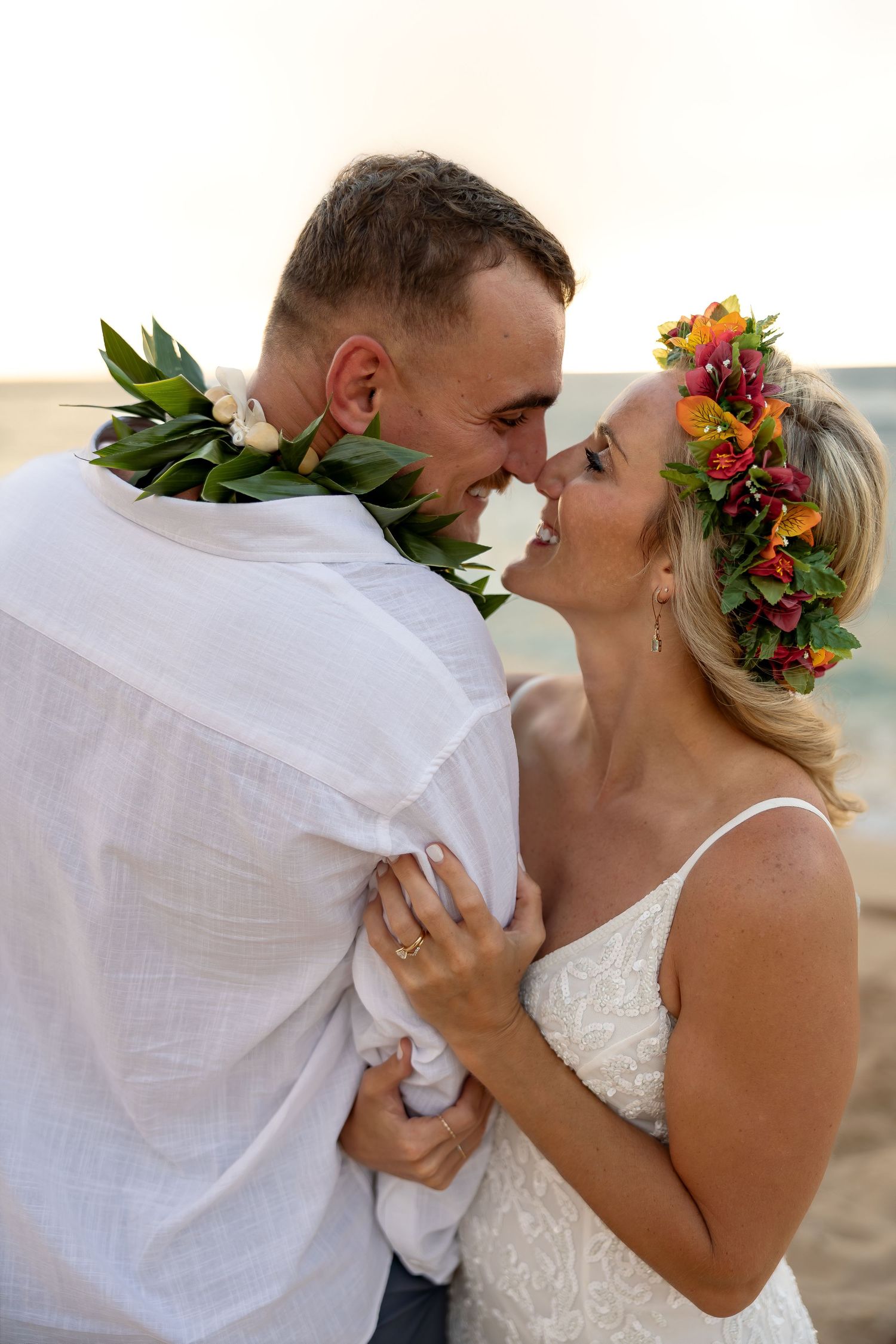 A smiling couple in floral leis nearly kiss during a beach wedding ceremony.