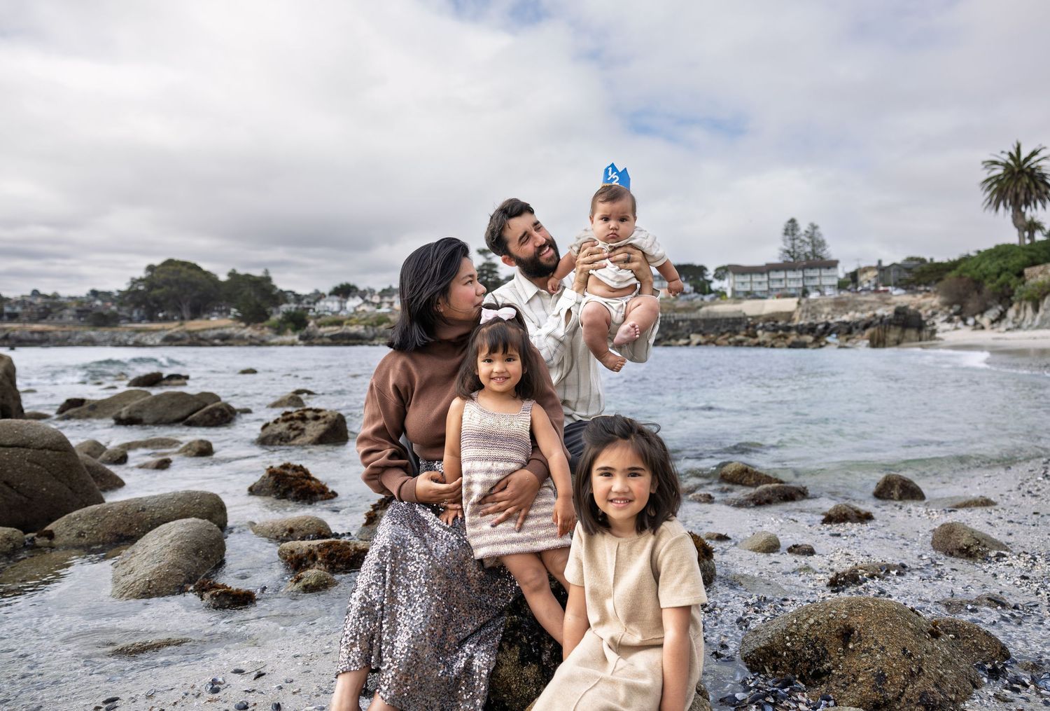 Family of five posing on rocky beach with ocean waves and cloudy sky in background.