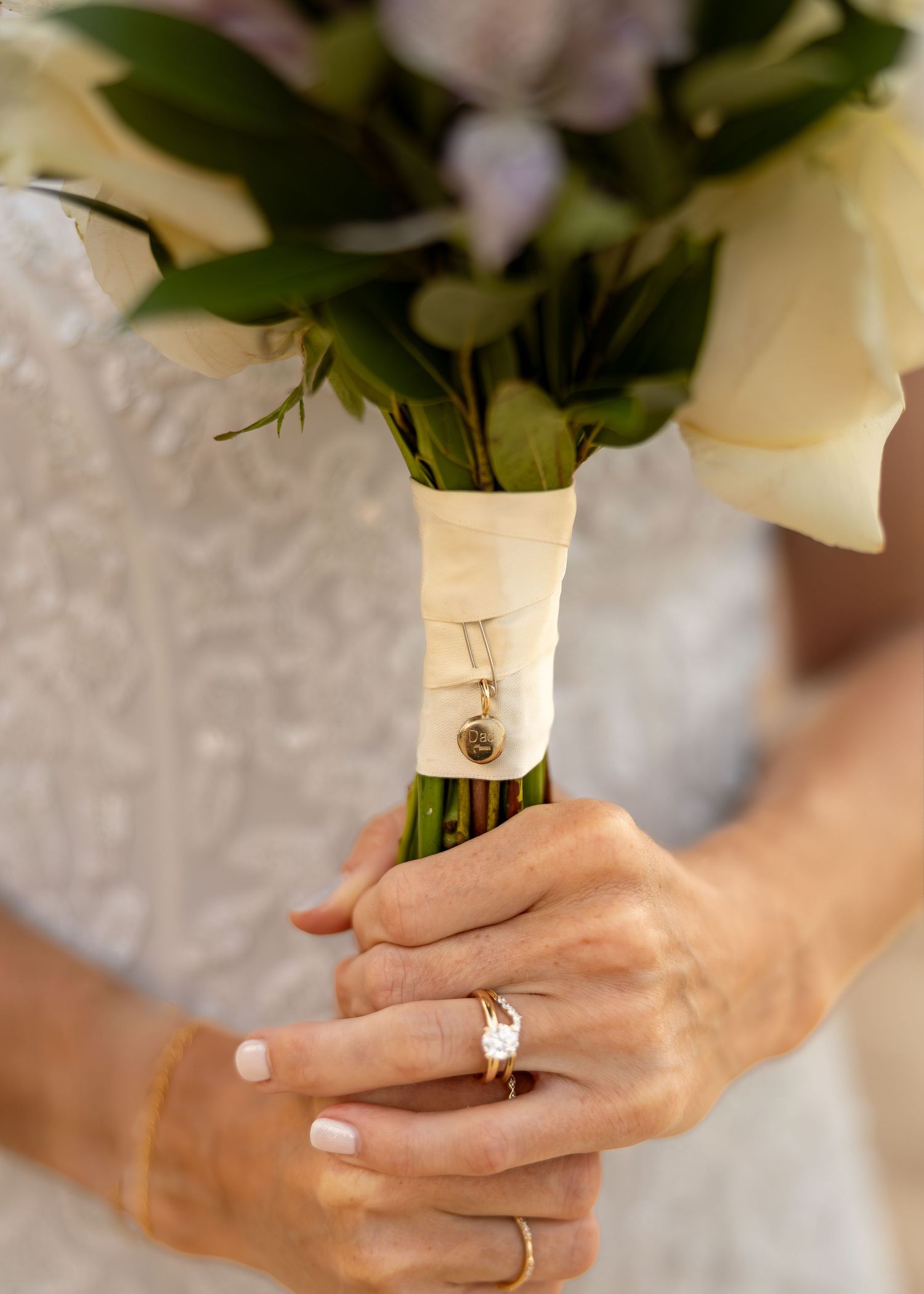 Bride holding white rose bouquet with ribbon and charm, wearing diamond engagement ring with French manicure.