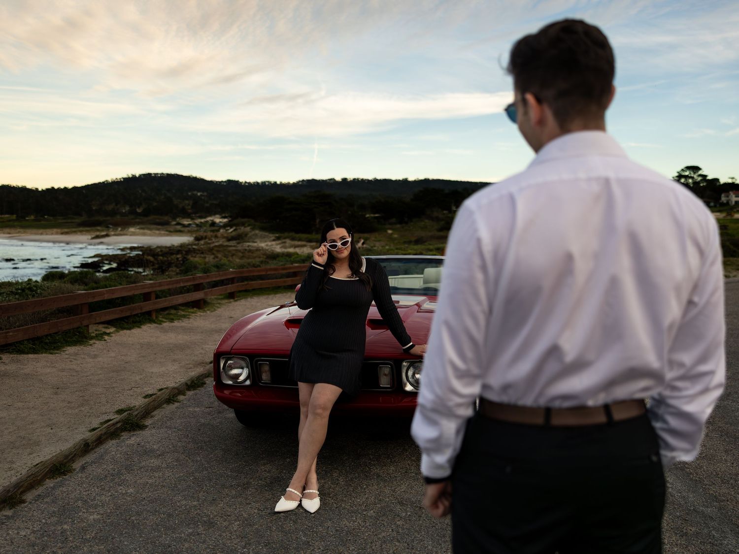 Woman posing by red convertible as man approaches on coastal road at sunset.