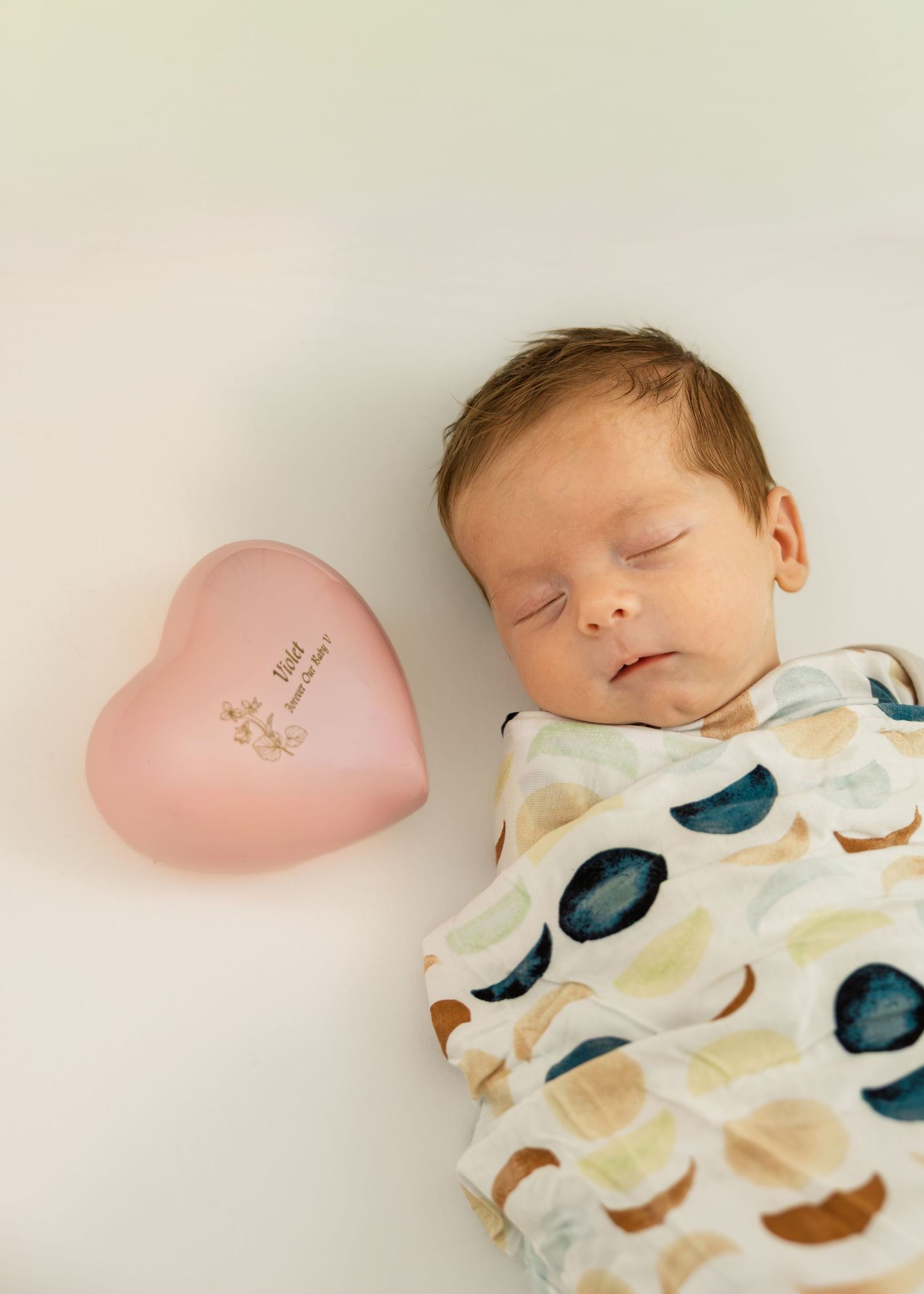 Sleeping newborn baby swaddled in a colorful blanket beside a pink heart-shaped keepsake box on a white background.