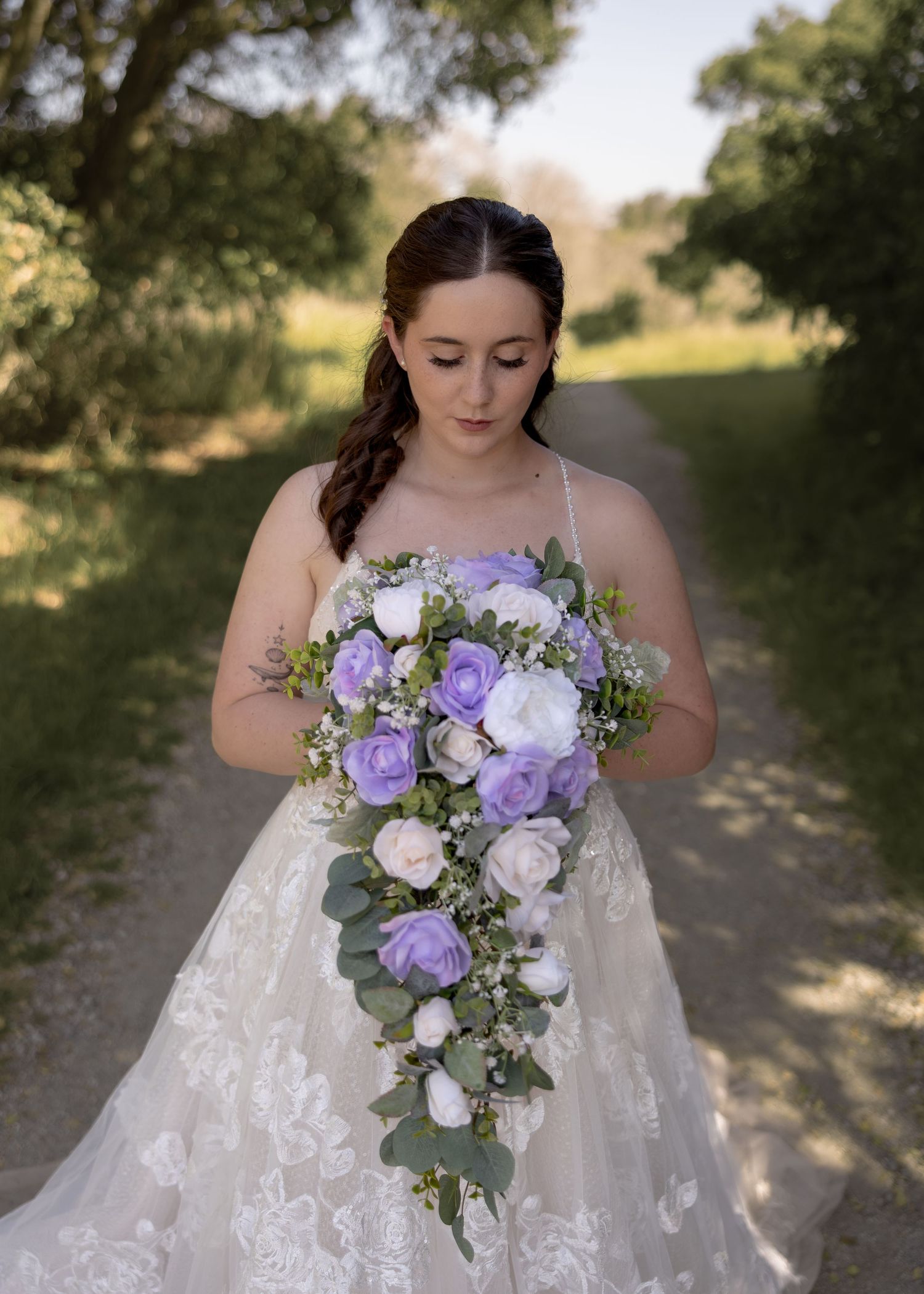 Bride in lace gown holds cascading purple and white floral bouquet on a tree-lined path.