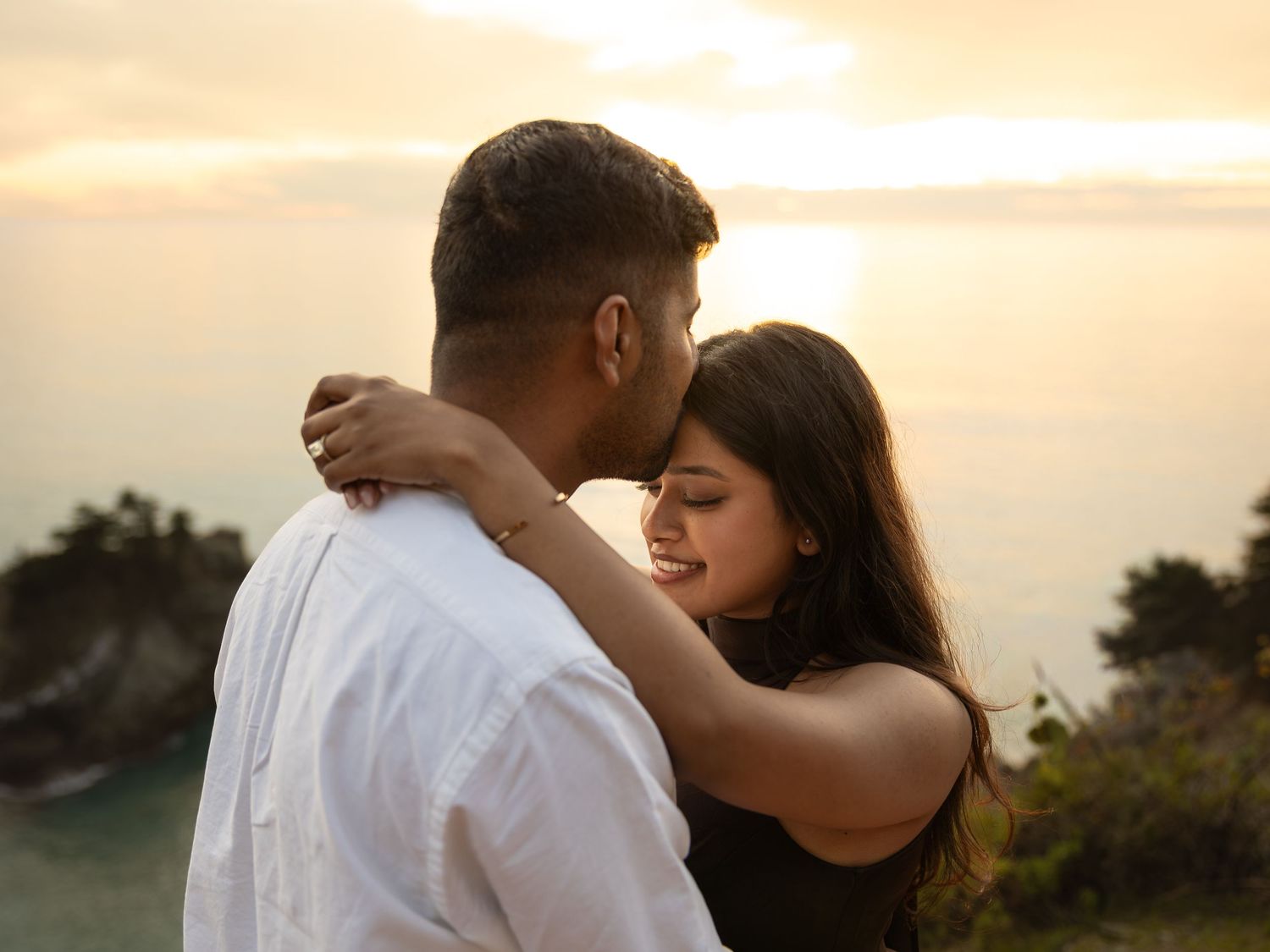 Romantic couple embracing at sunset with ocean views, woman smiling as man kisses her forehead outdoors.