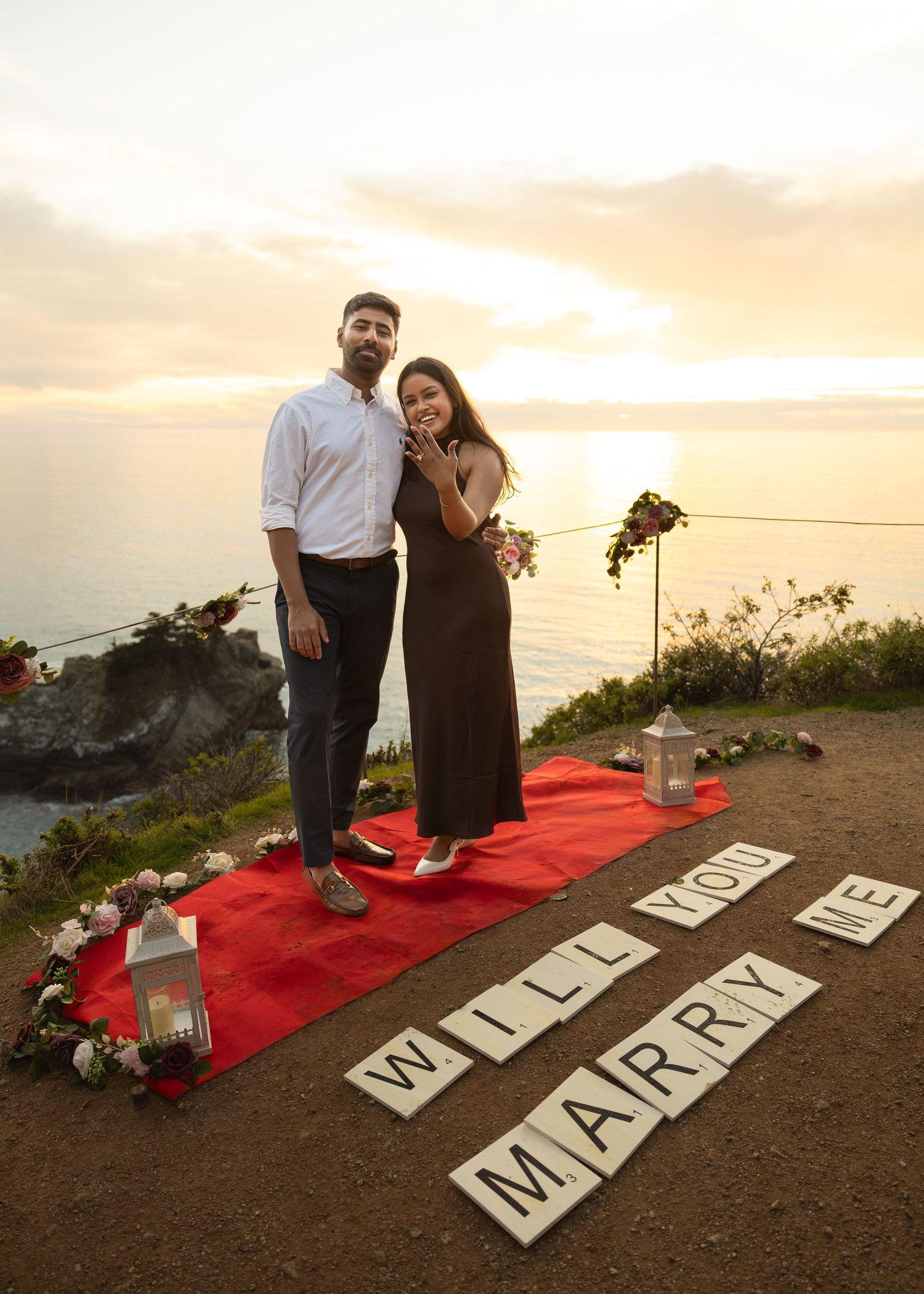 Couple poses at sunset cliffside proposal with 'Will You Marry Me' Scrabble tiles on red carpet.