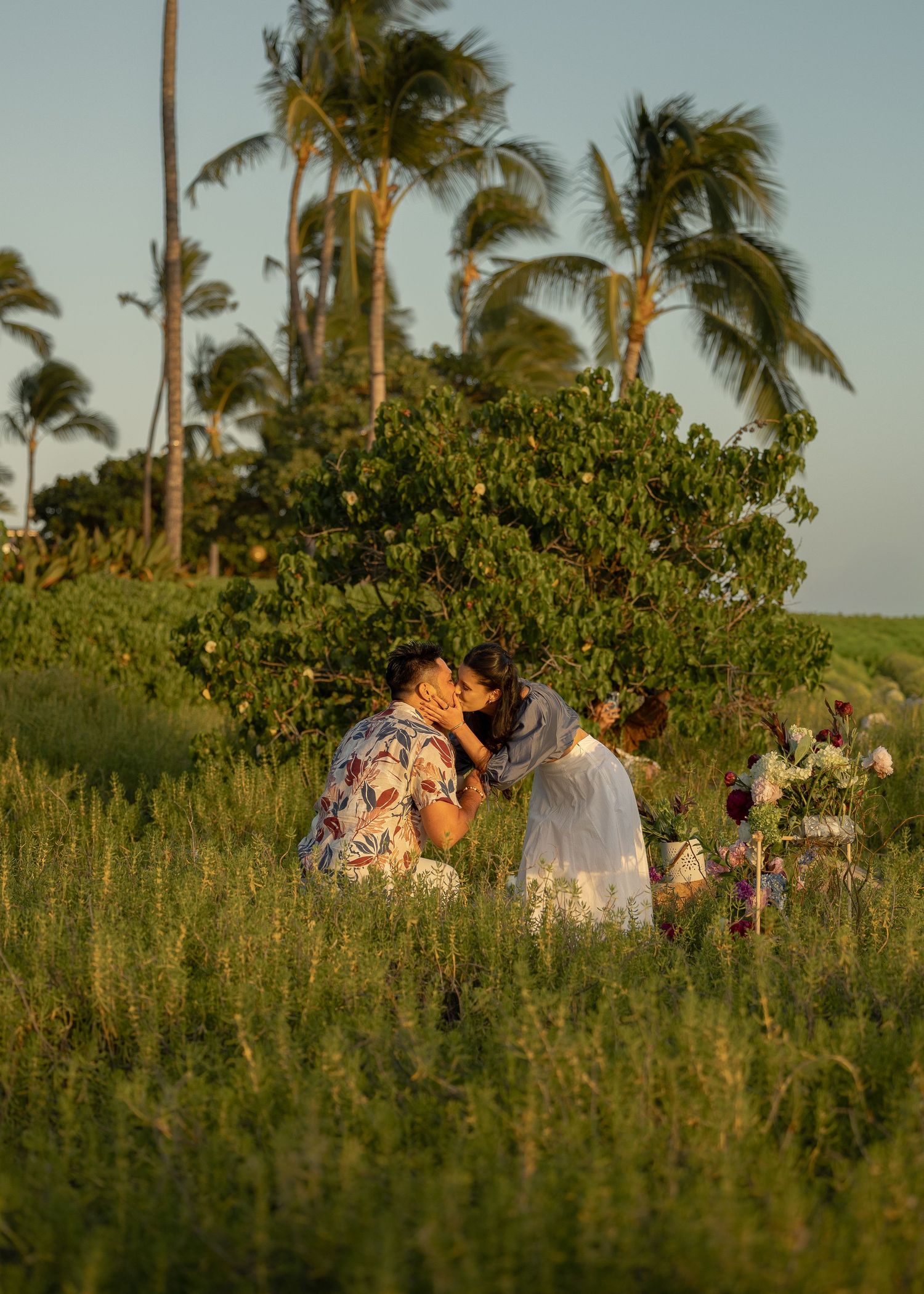 Couple sharing a romantic kiss in a lush green field with tropical palm trees at golden hour.