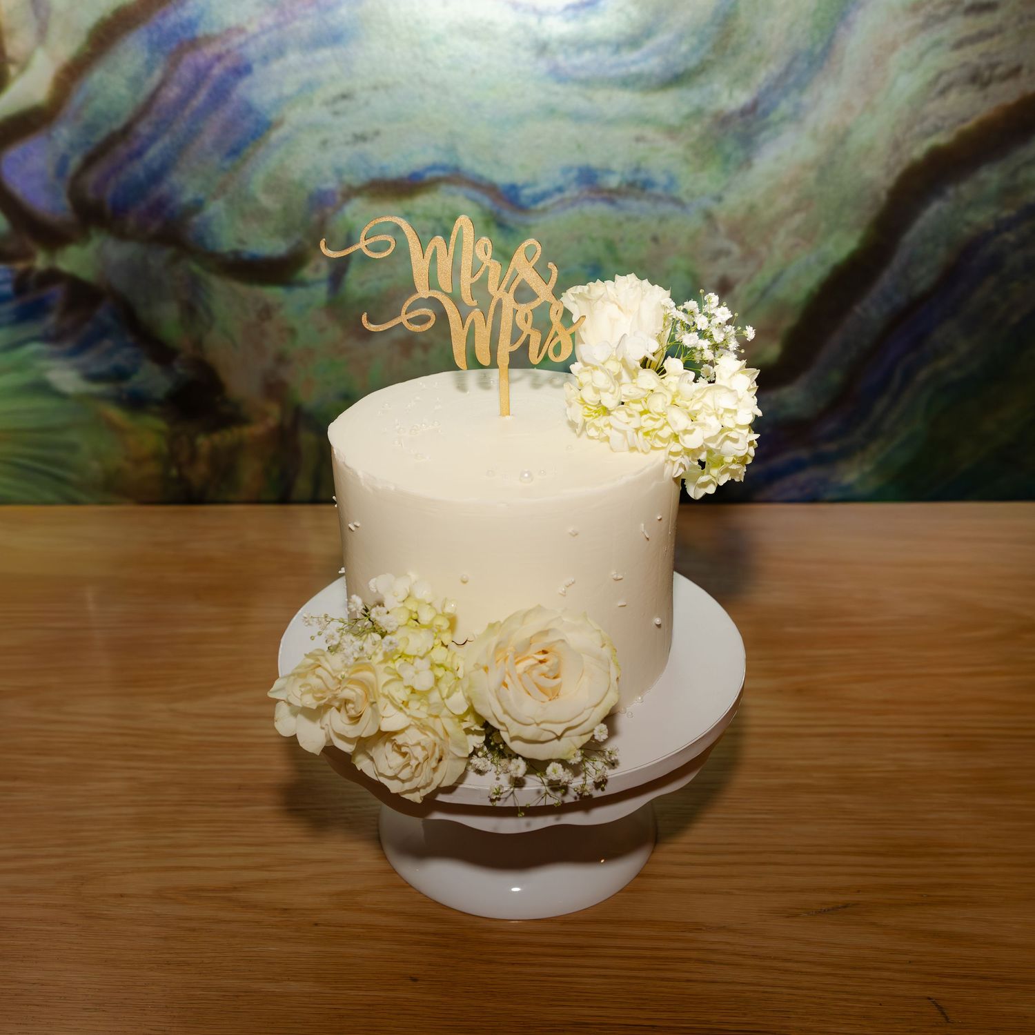 Elegant white wedding cake with cream roses, hydrangeas, and a gold Mrs. & Mrs. topper on a white cake stand.