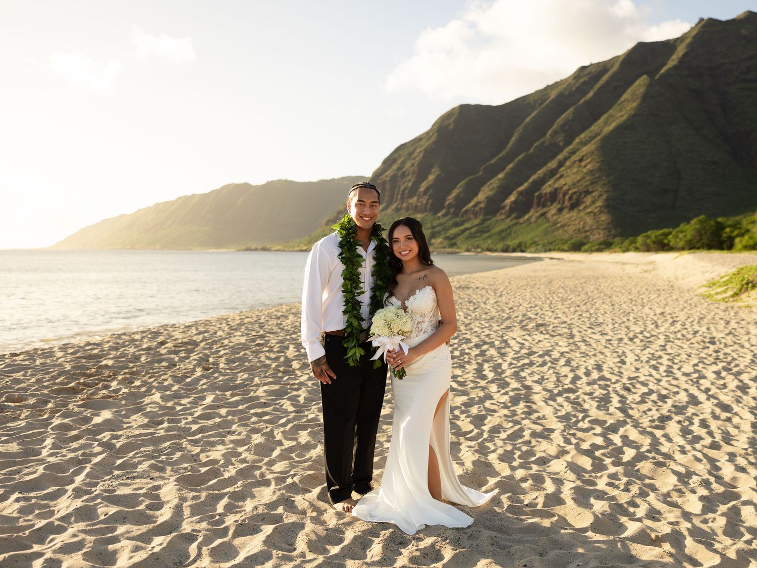 Couple in wedding attire posing on a sandy Hawaiian beach with lush green mountains and golden sunset in the background.