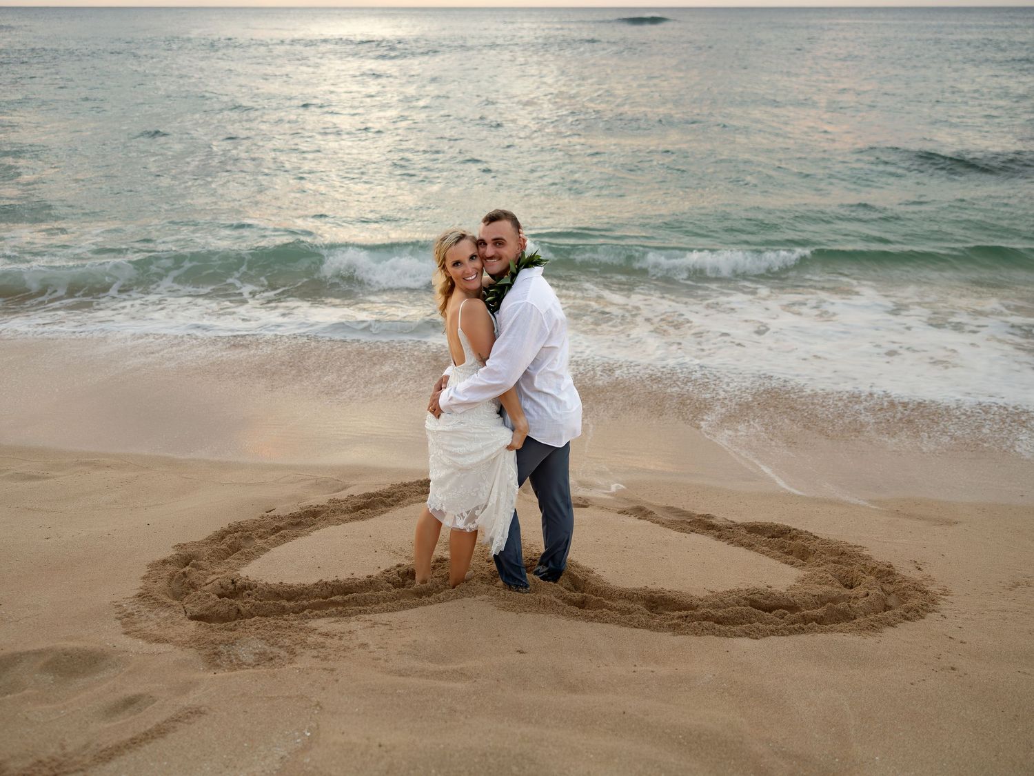 Couple embracing inside a heart drawn in sand on a beach at sunset, romantic seaside engagement photo.