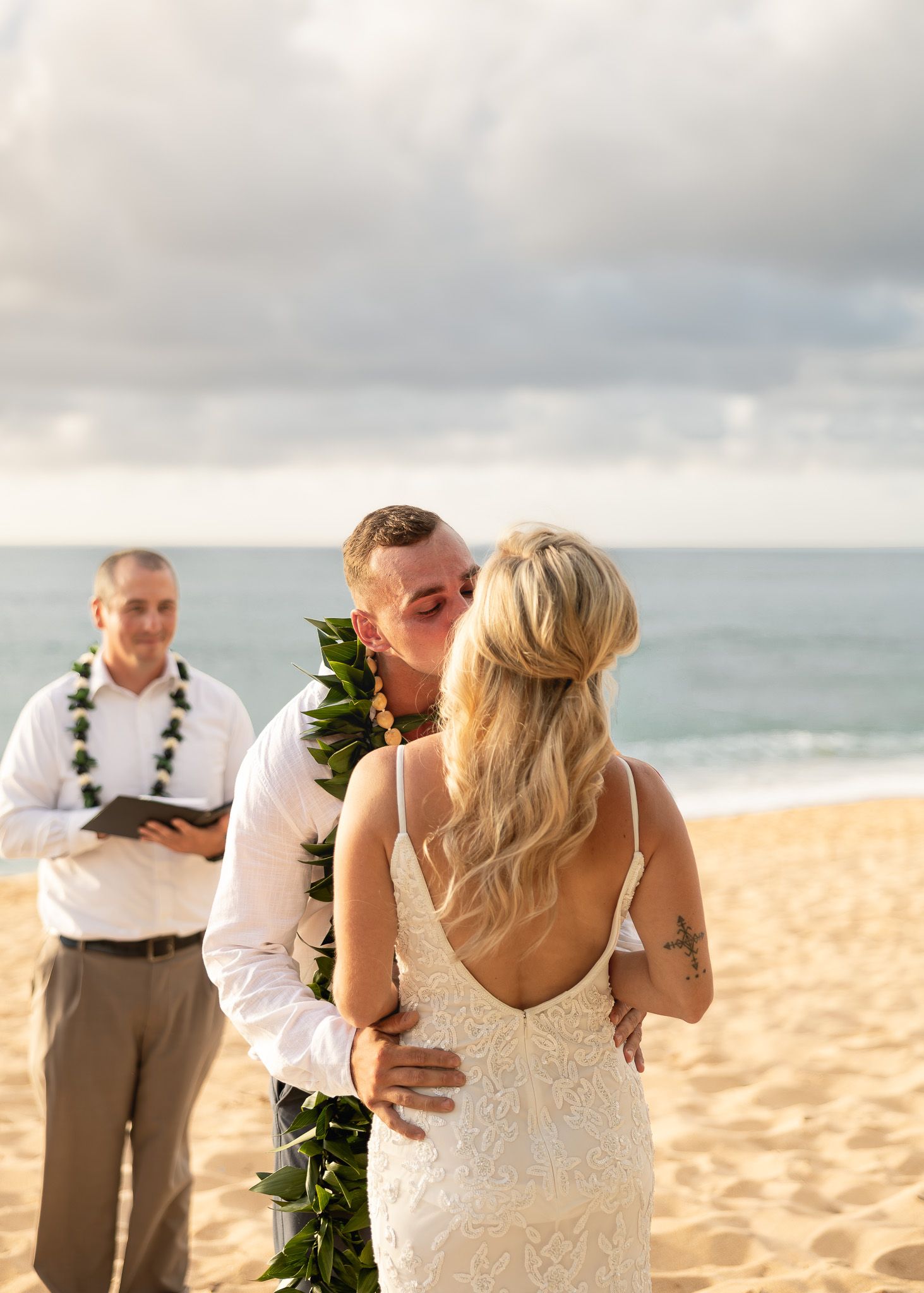 Couple sharing first kiss on sandy beach wedding ceremony in Hawaii with officiant wearing lei in background.