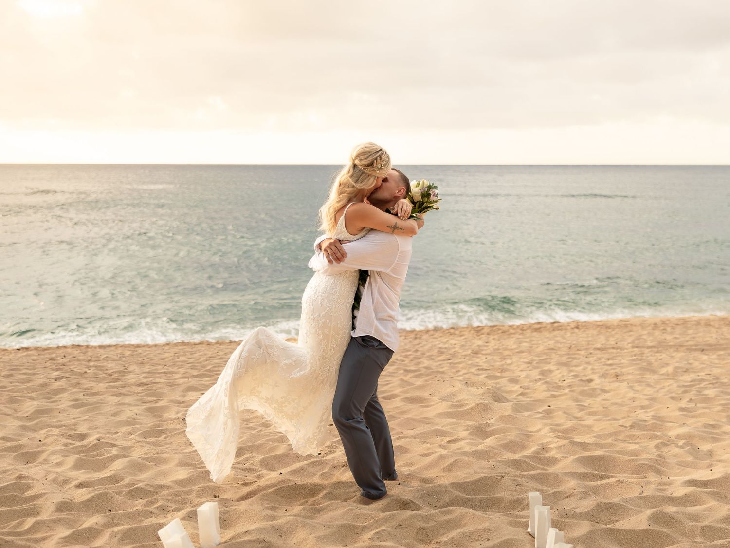 Groom lifting bride in wedding dress on sandy beach with ocean waves in background at sunset.