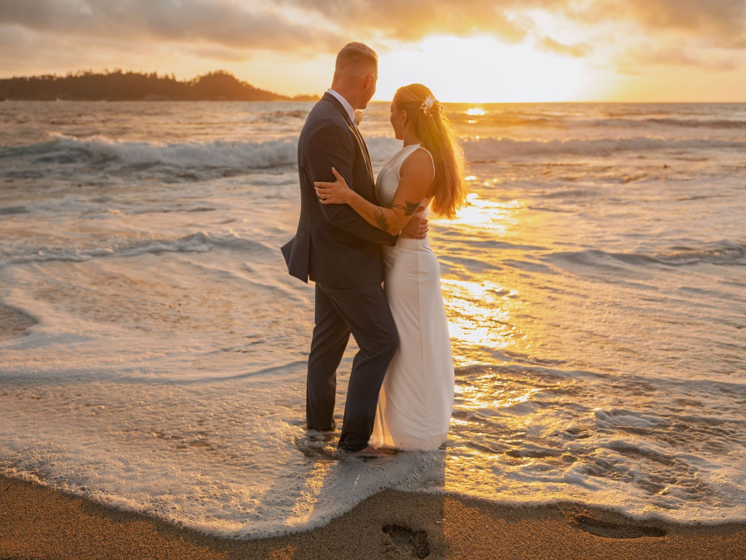 Newlywed couple embracing at waters edge on beach during stunning golden sunset ceremony.