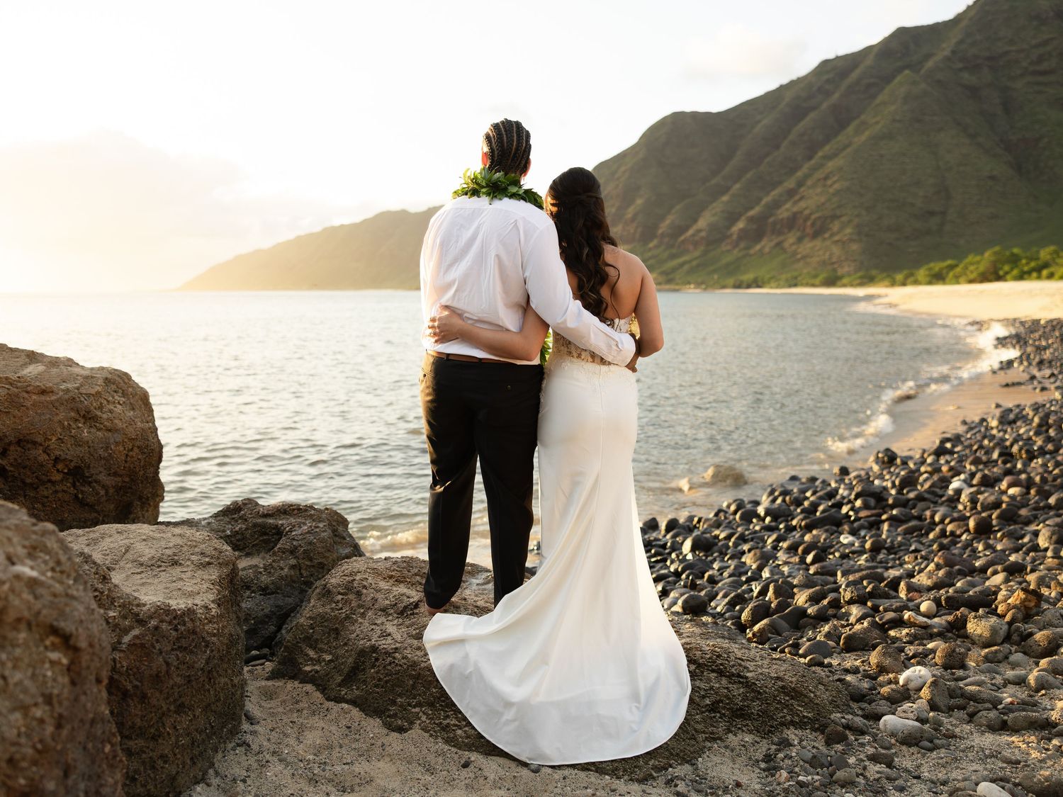 Newlywed couple embraces overlooking scenic Hawaiian coastline with mountains at sunset.