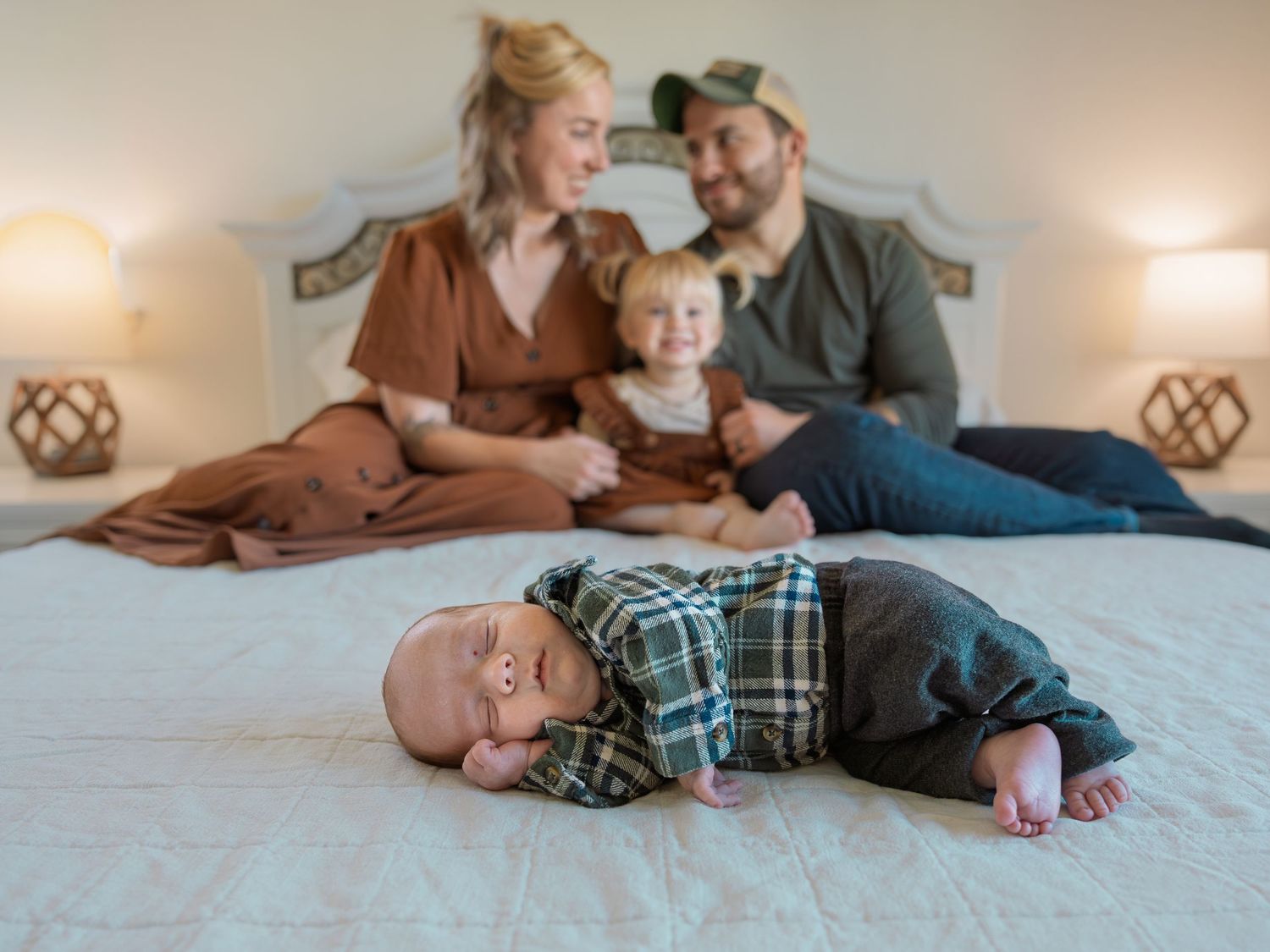 A newborn baby lies on a white bed while parents and toddler sit together in a warm family portrait.