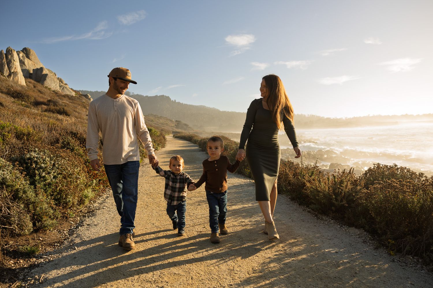Family of four holding hands walking along a scenic coastal path with ocean views and golden sunlight.