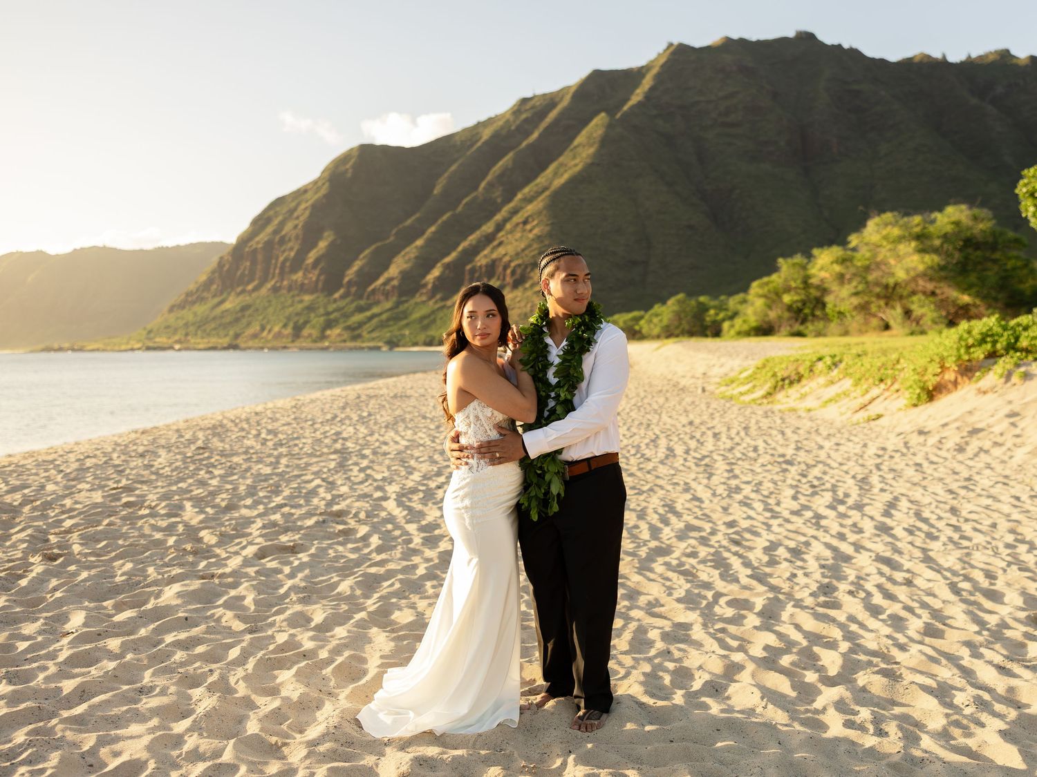 Couple in wedding attire poses on sandy Hawaiian beach with lush green mountains and golden sunset light behind them.